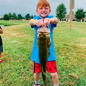 Child holding a large fish outdoors on grass, smiling, with trees and a building in the background.
