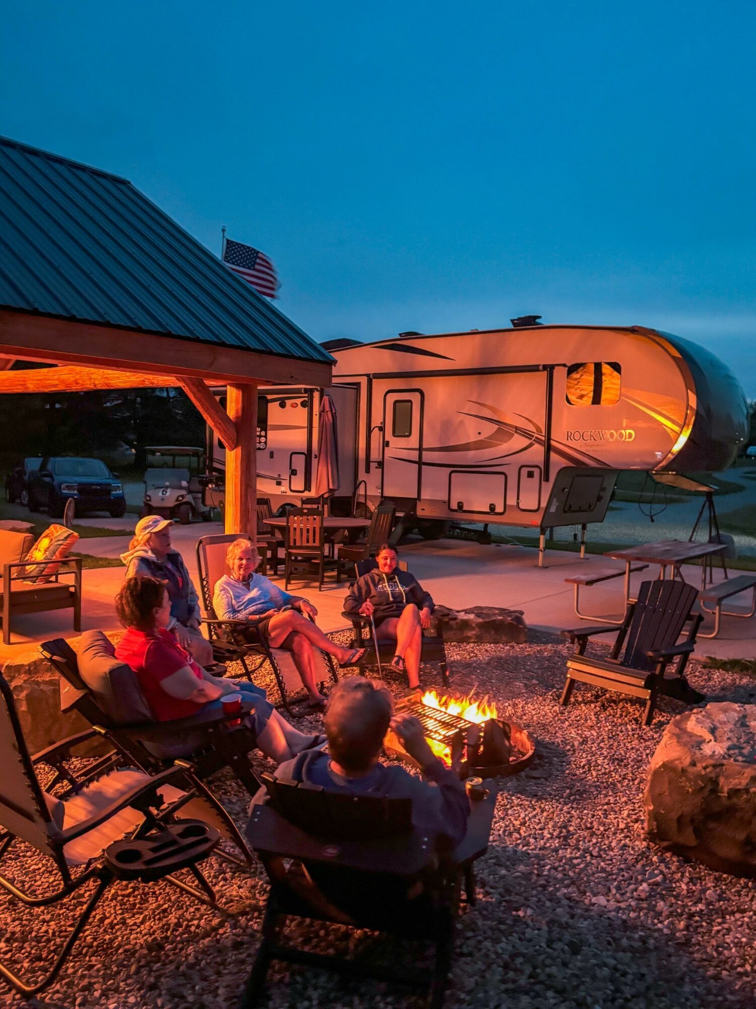 People sitting around a campfire outside a building with a rocket in the background during evening.