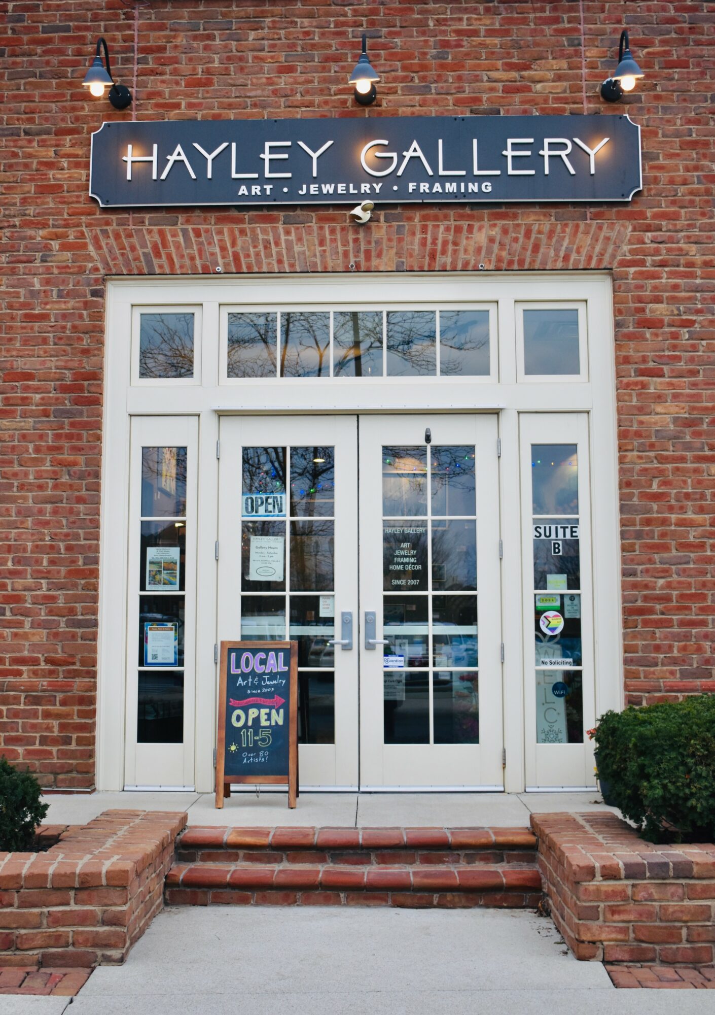 Brick building entrance with glass double doors, sign reading 'Hayley Gallery,' and an A-frame sign outside, with bushes on the side.