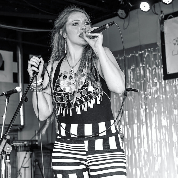 Woman singing into a microphone on stage with a curtain backdrop, wearing striped pants and jewelry.