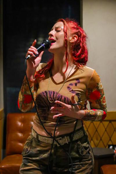 Woman with red hair singing into a microphone, wearing a colorful top, in a room with a brown chair and yellow wall.