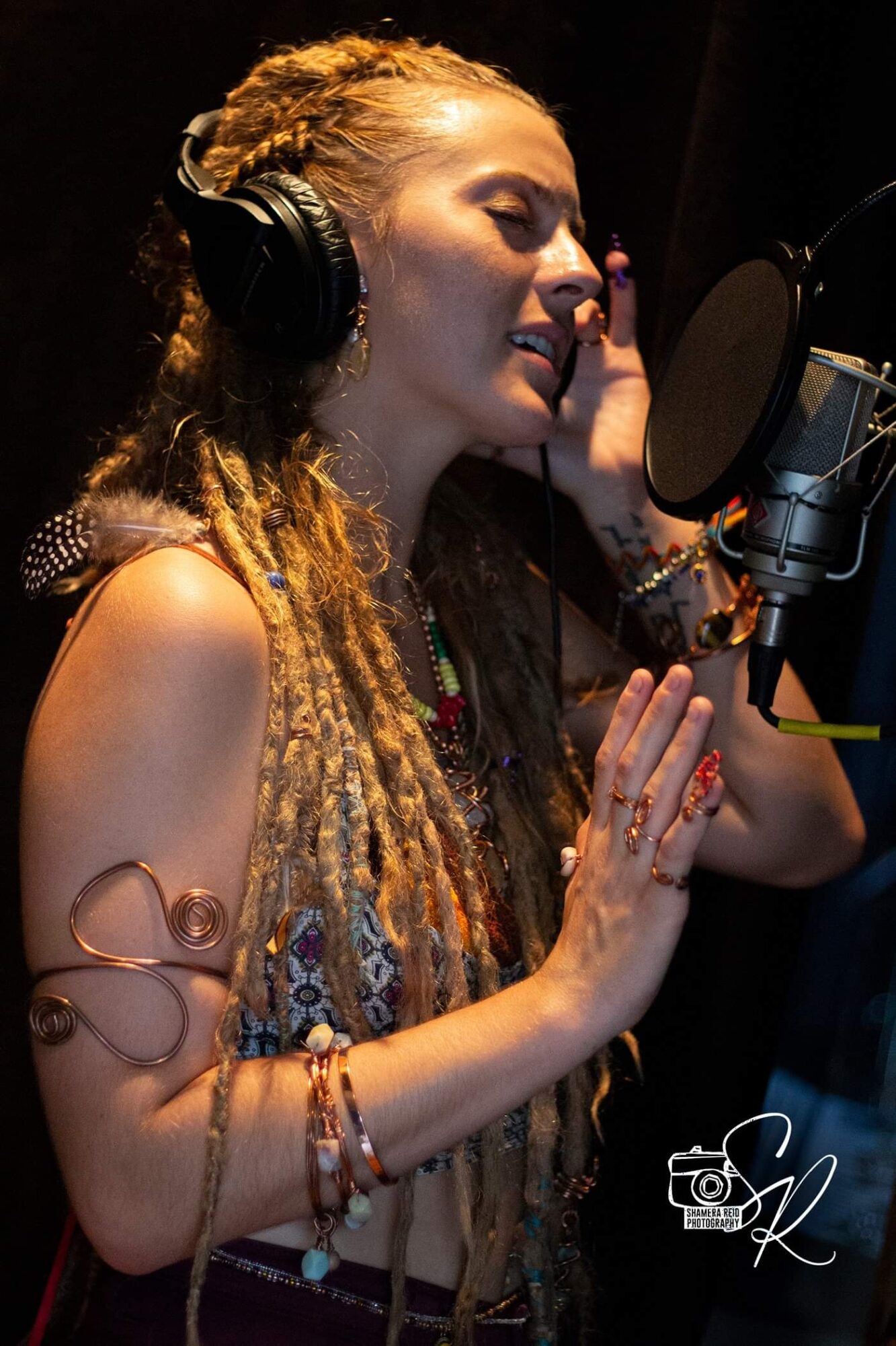 Woman with long dreadlocks singing into a microphone, wearing headphones and jewelry, in a recording studio.