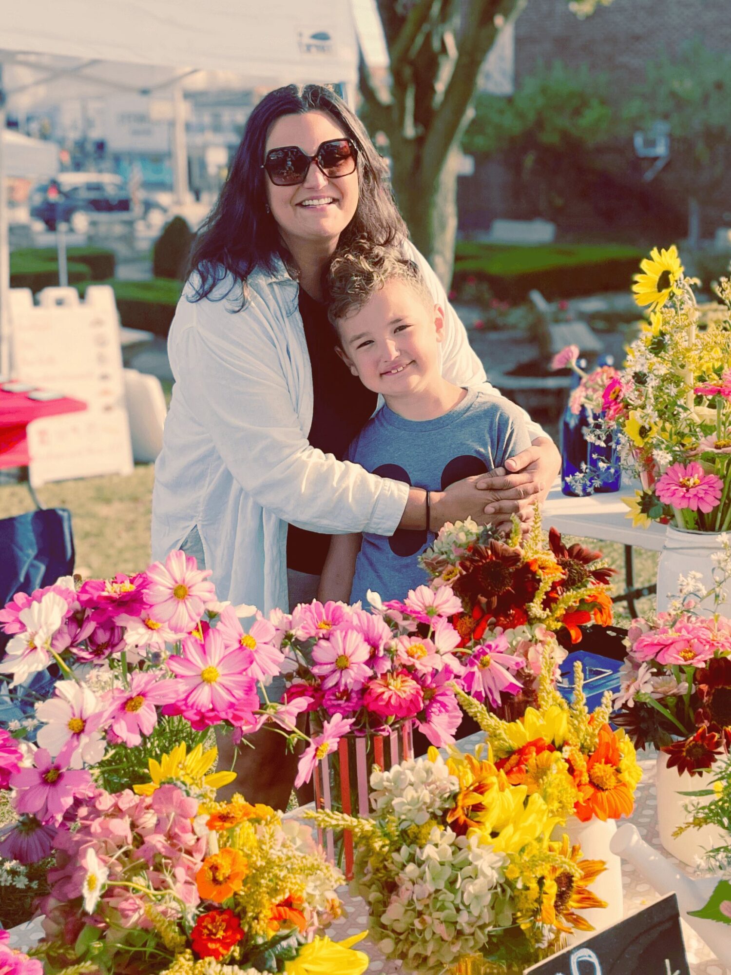 Woman and child smiling at a flower stand outdoors with colorful flowers and trees in background.