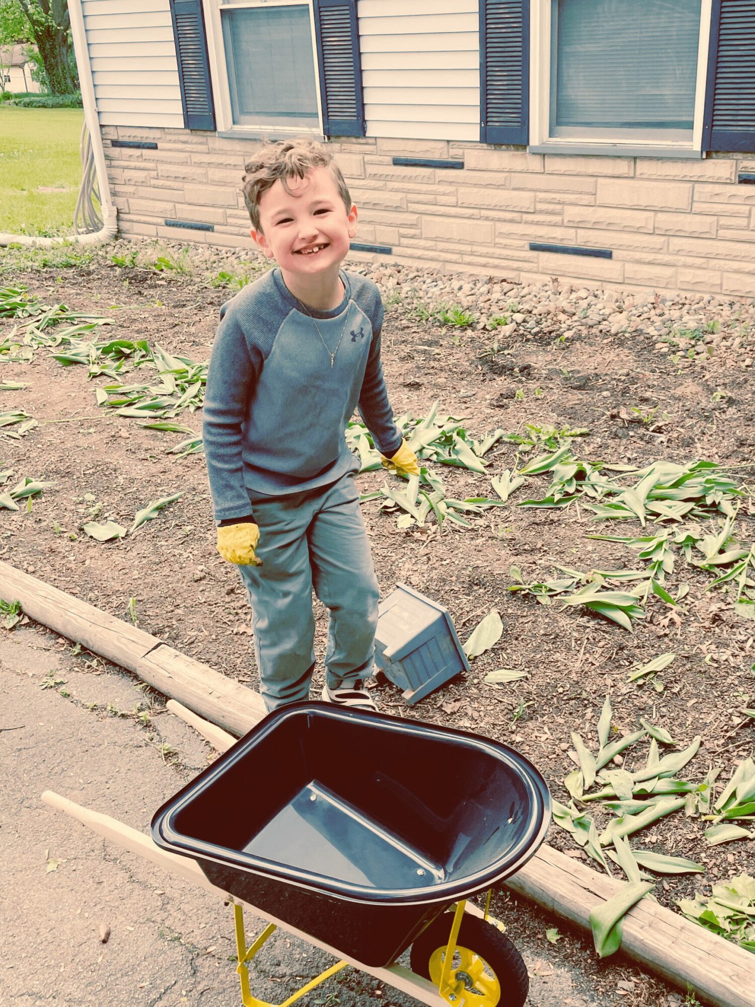 Young boy smiling in a garden, holding gardening tools, with a house in the background.