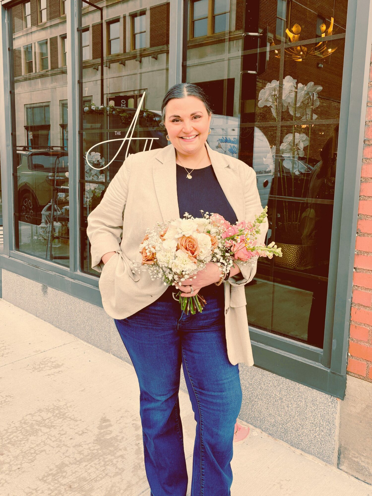 Woman holding a bouquet of flowers standing outside a building with large glass windows, smiling, wearing a beige blazer and blue jeans.