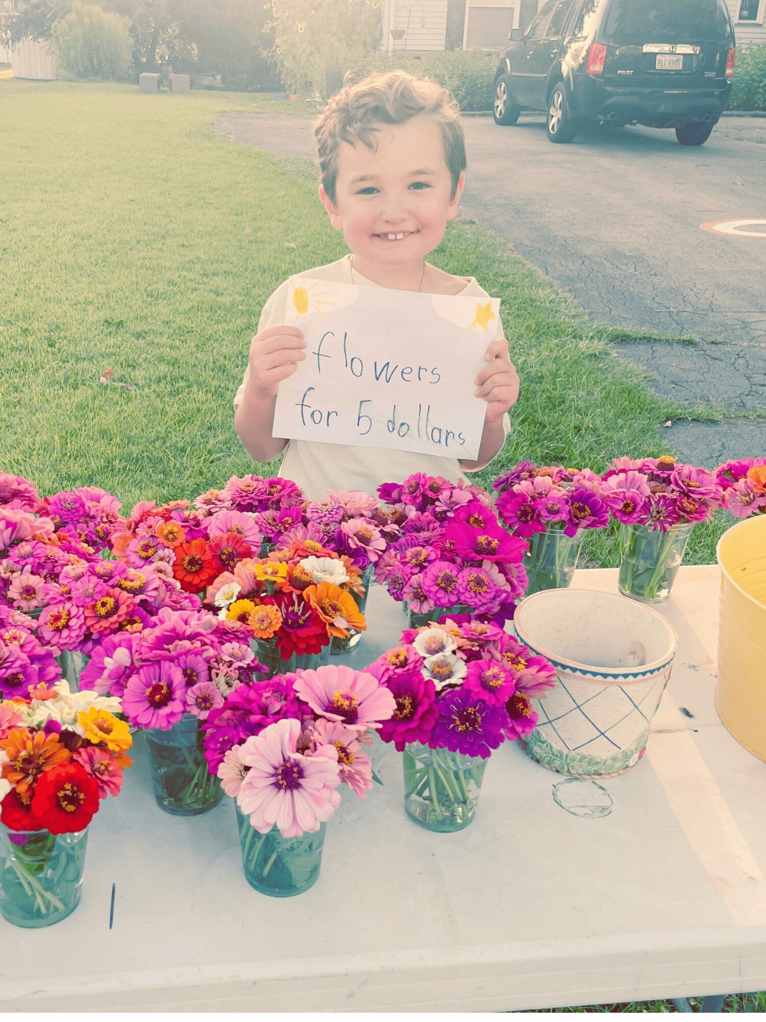 Smiling young boy holding a sign that reads 'flowers for 5 dollars' stands behind a table with colorful flowers in small vases outdoors.