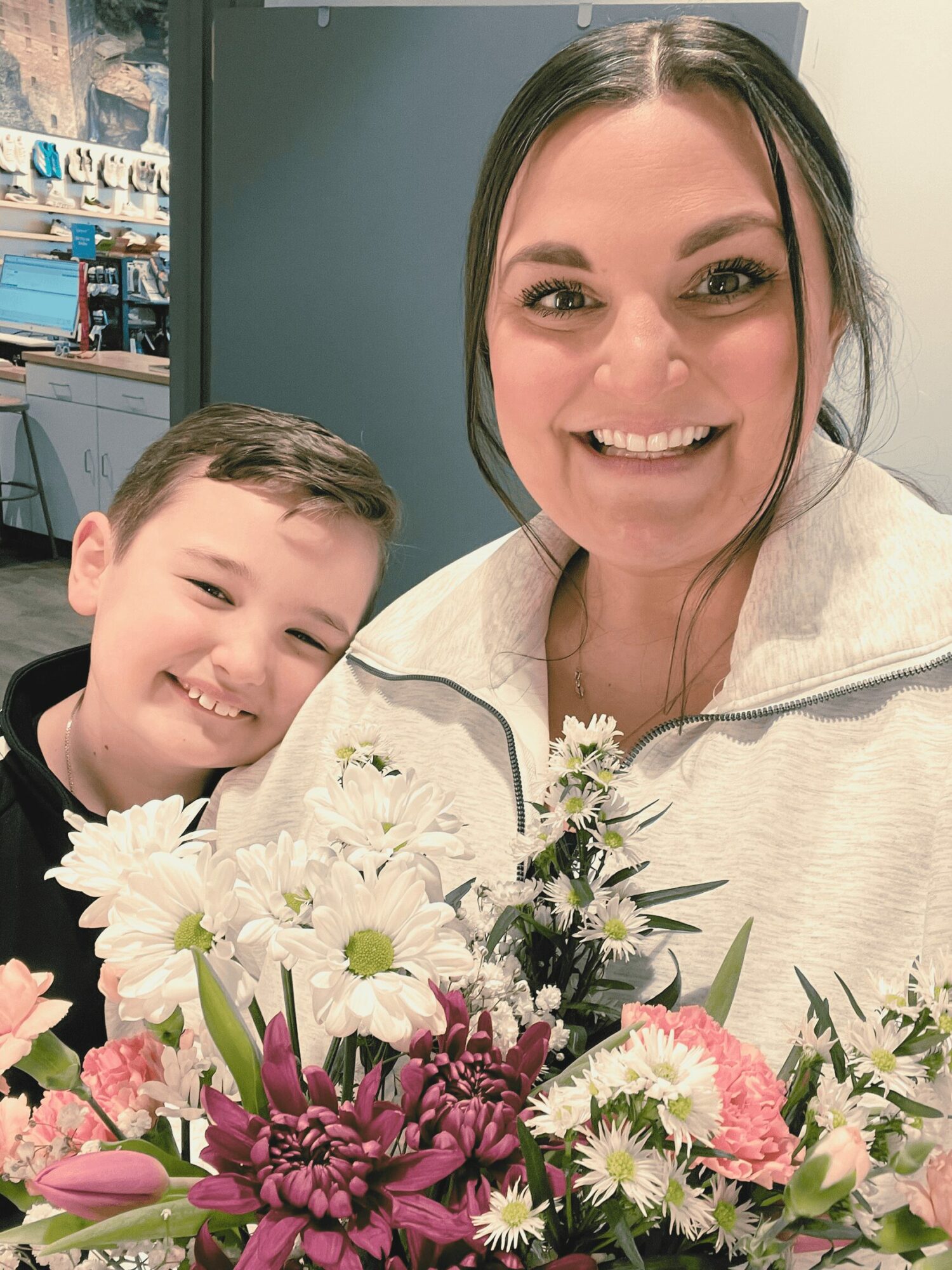 A woman and a young boy smiling, holding a bouquet of flowers, indoors with shelves in the background.