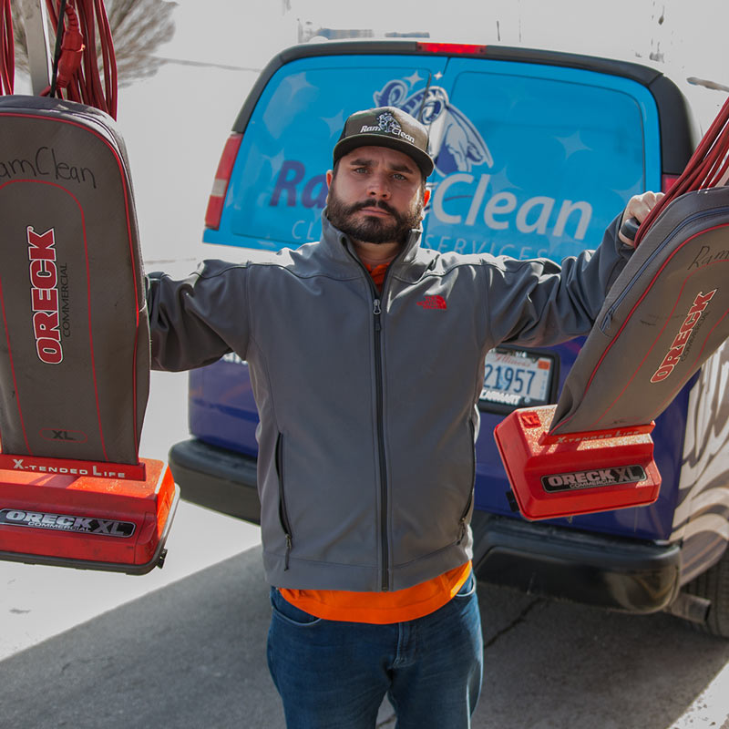 Man holding two ORECK vacuum cleaners outdoors, with a blue vehicle in the background.