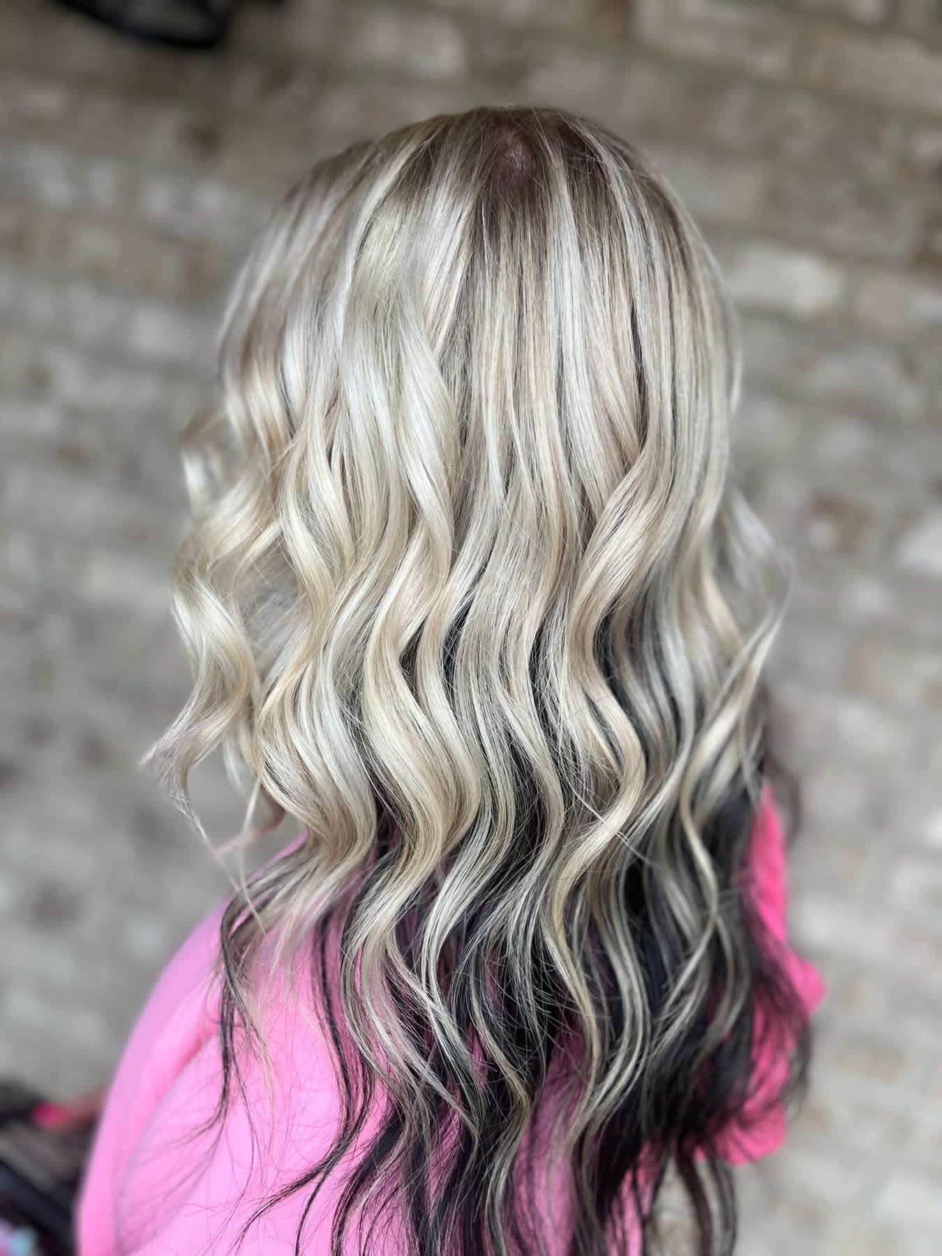 Back of a person with long, wavy, blonde hair, wearing a pink top, against a brick wall background.