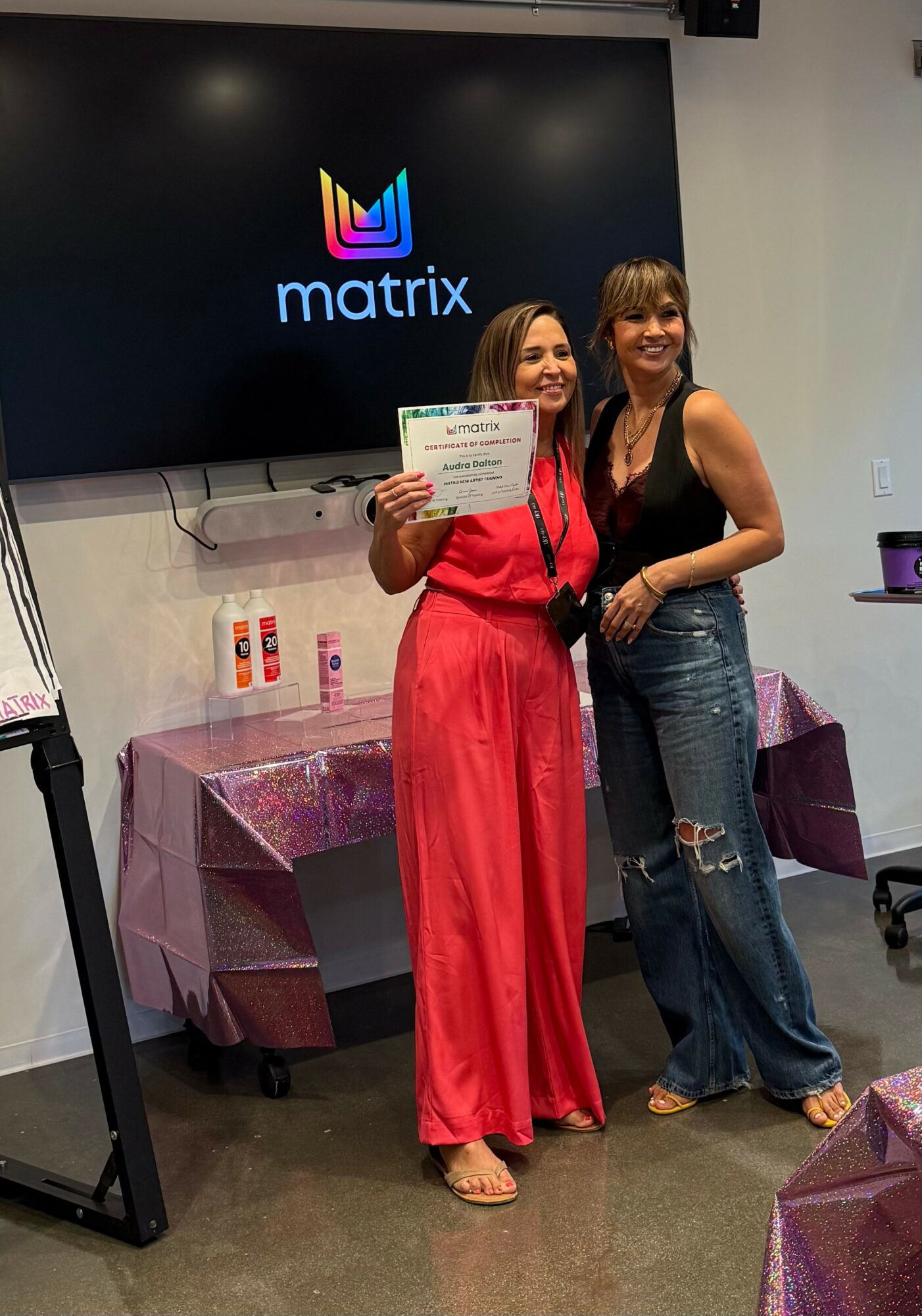 Two women standing together, one holding a certificate, smiling in front of a large screen with a logo, in a room with tables covered in pink cloths.