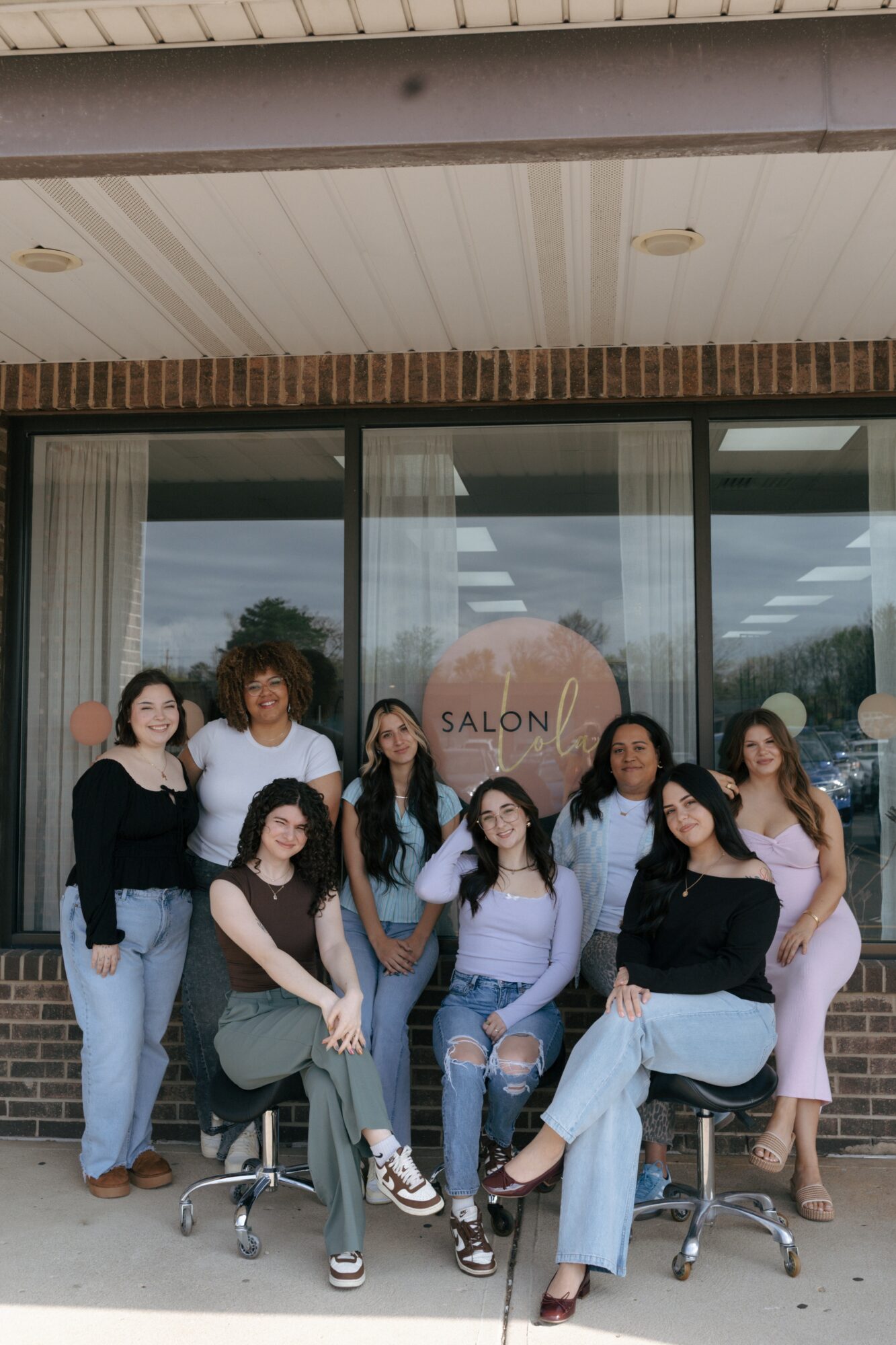 Group of nine women posing outside a salon, some sitting and some standing, in front of glass windows.