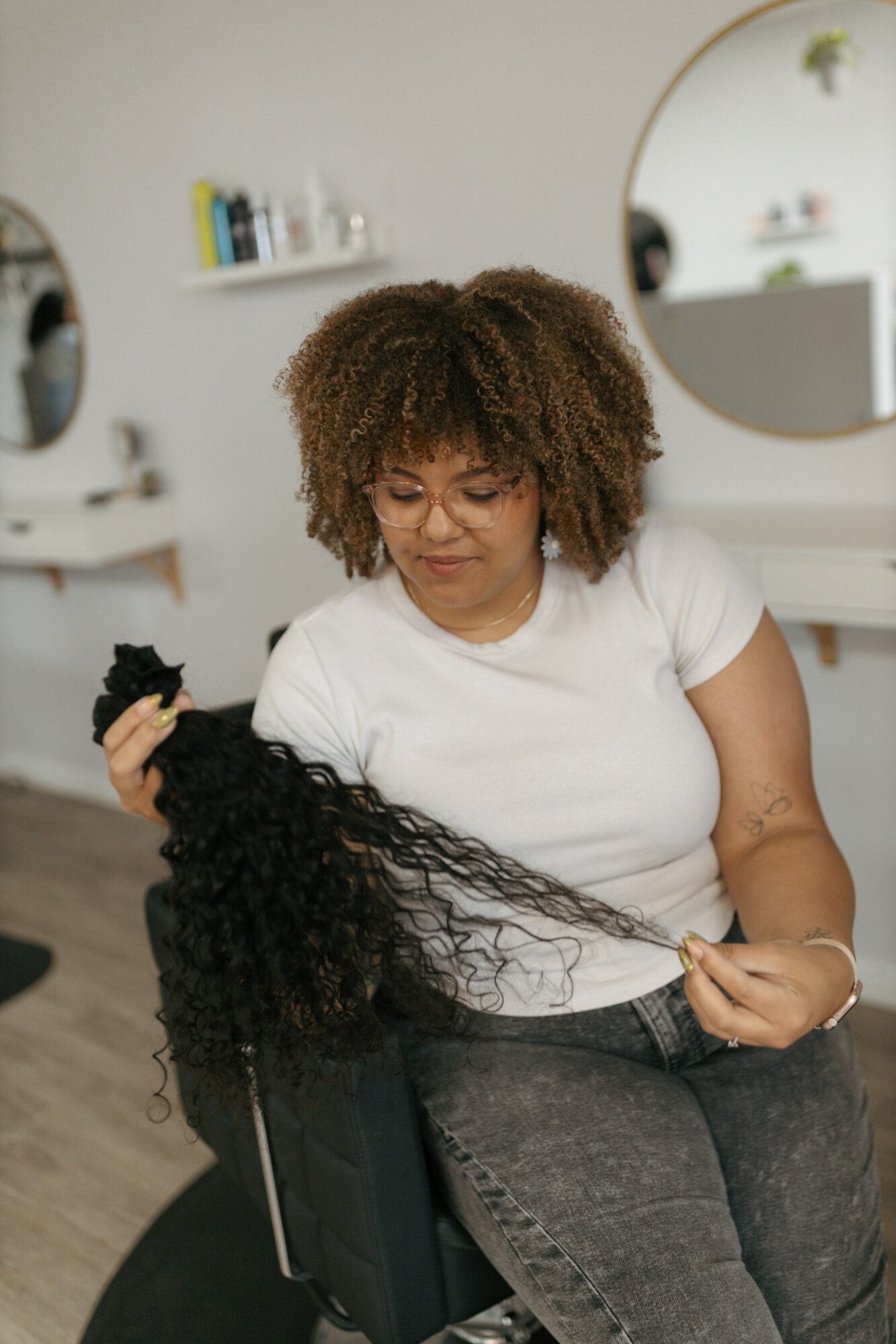 Woman with curly hair holding a long, black, curly wig in a room with a mirror and shelves.