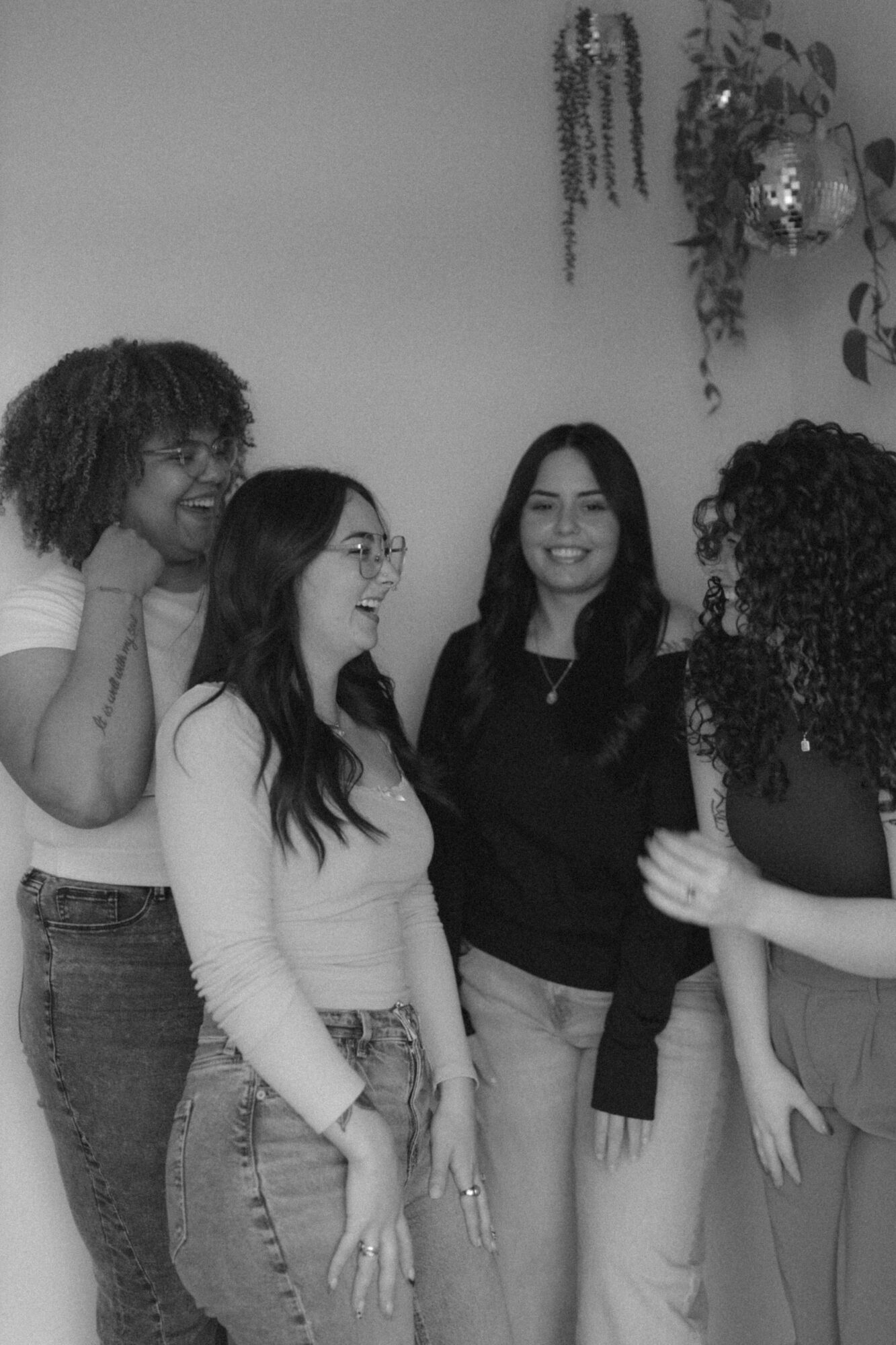 Four women standing together, smiling and talking indoors, with hanging plants and decorations on the wall behind them.