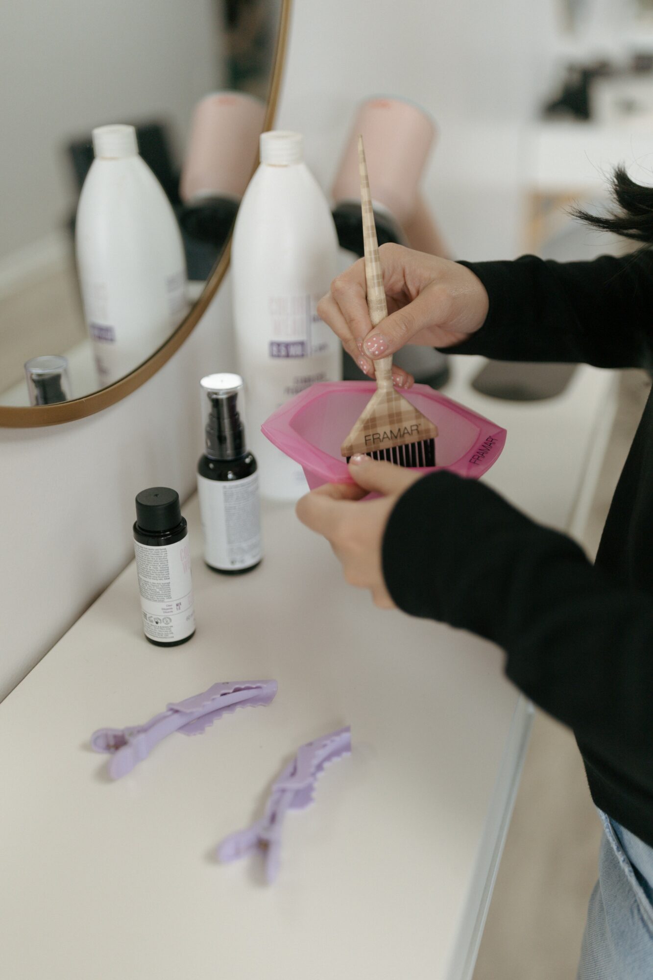 Person holding a makeup brush over a pink container on a bathroom counter with bottles and brushes, mirror in background.