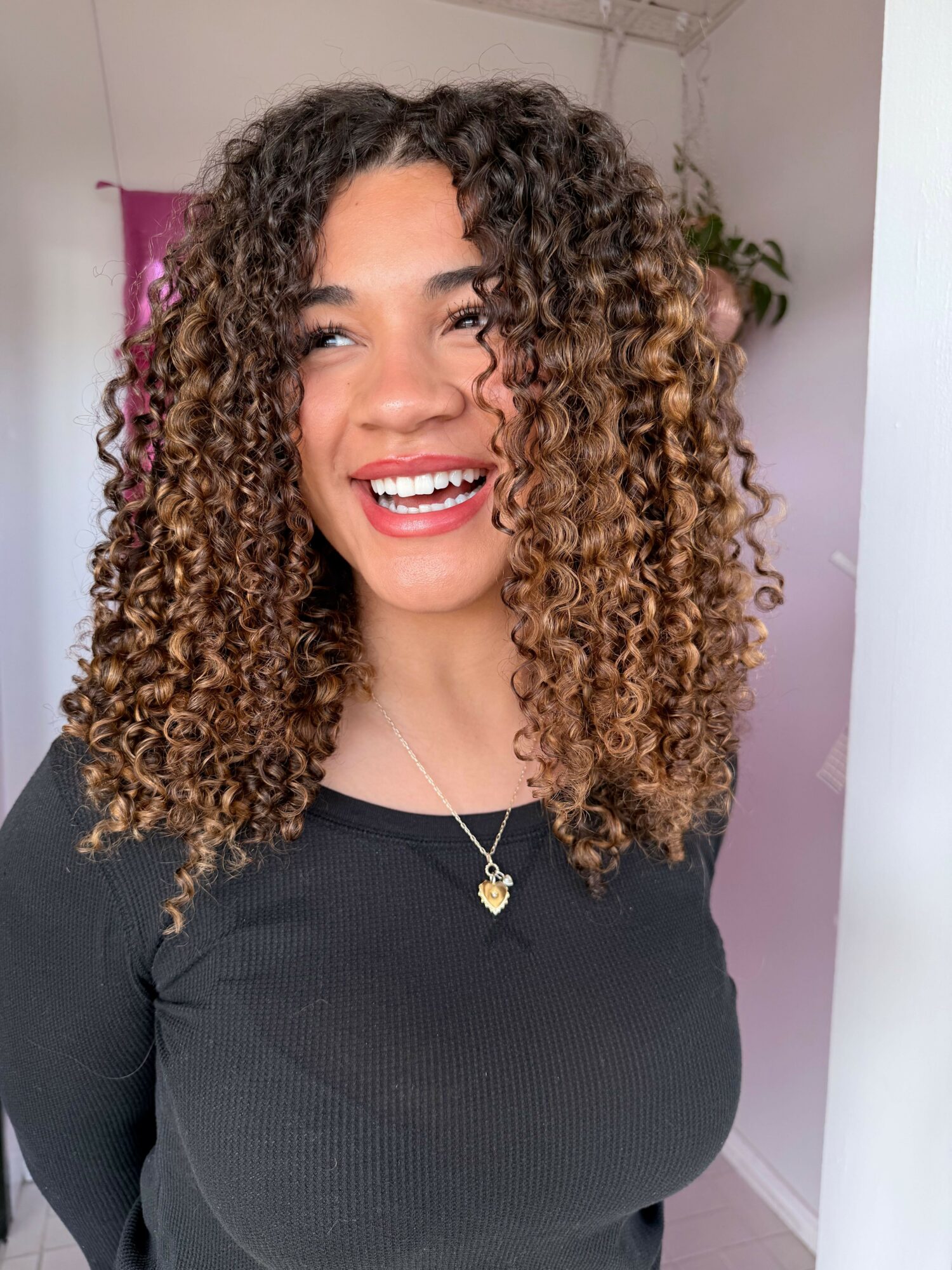 Smiling woman with curly hair wearing a black top and gold necklace, standing indoors.