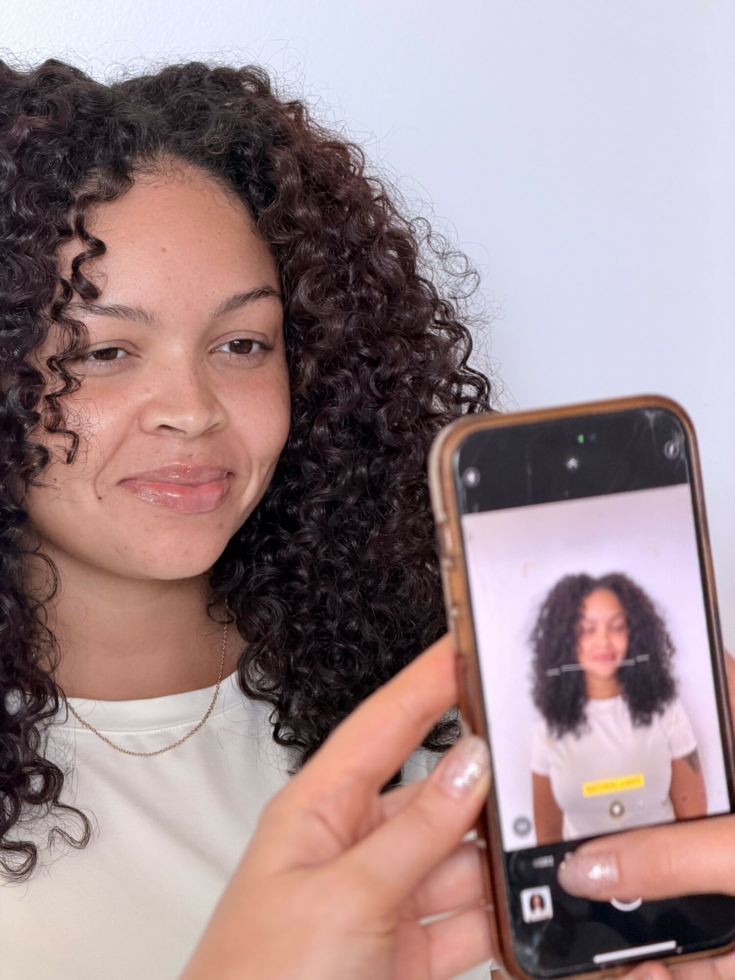 Person with curly hair taking a selfie, holding a smartphone, with a neutral background.
