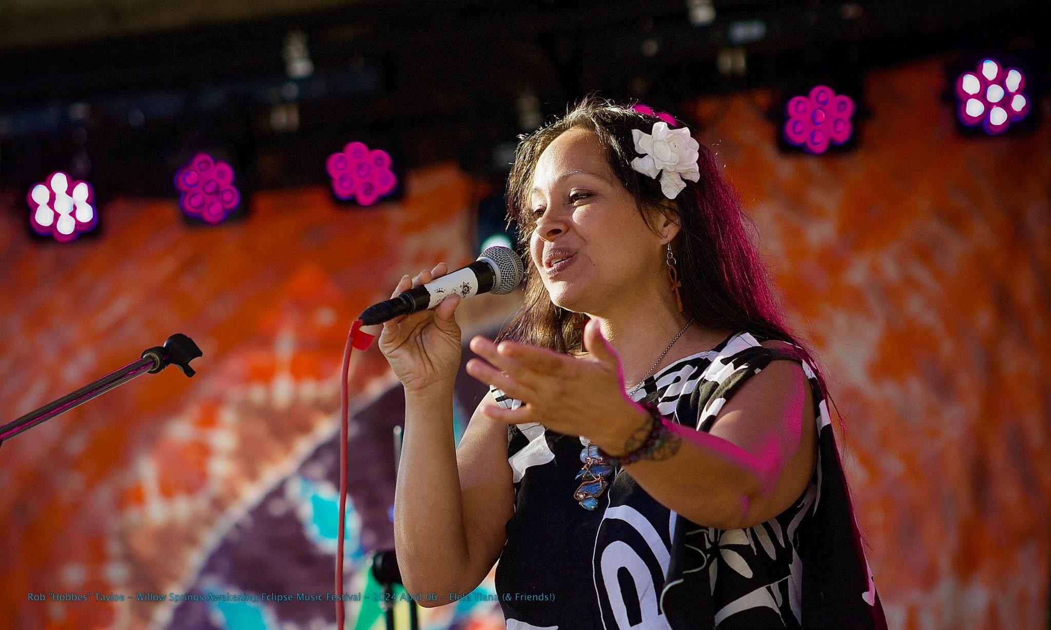 Young woman singing into a microphone on stage with colorful background and purple stage lights.