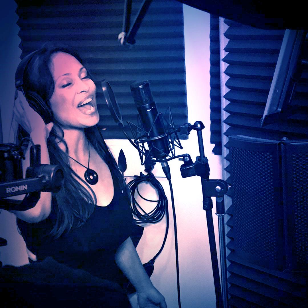 Woman singing into a microphone in a recording studio with soundproofing panels, wearing headphones and a black top.