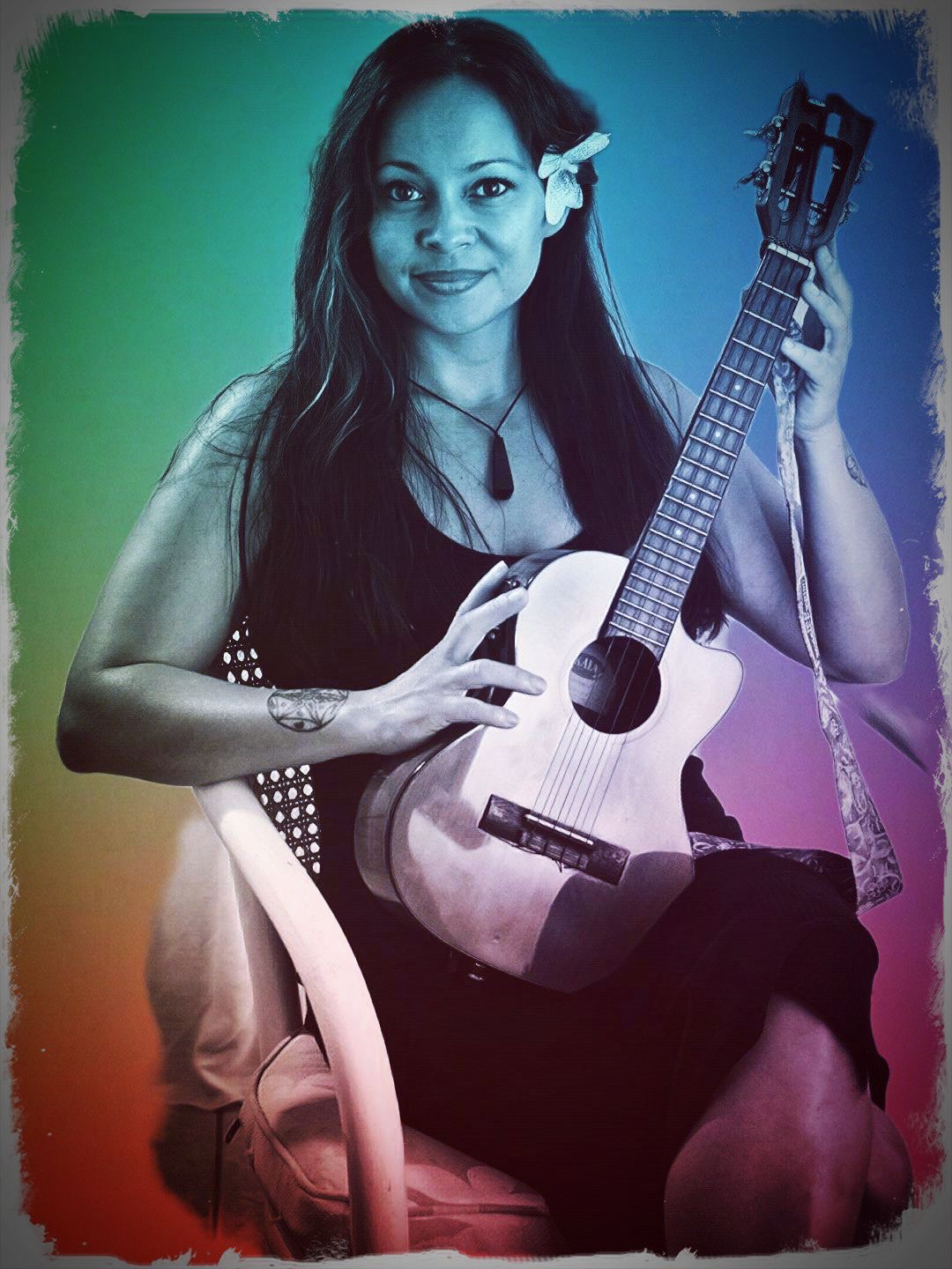 Woman with long hair holding a guitar, sitting on a chair, with a flower in her hair, against a colorful background.