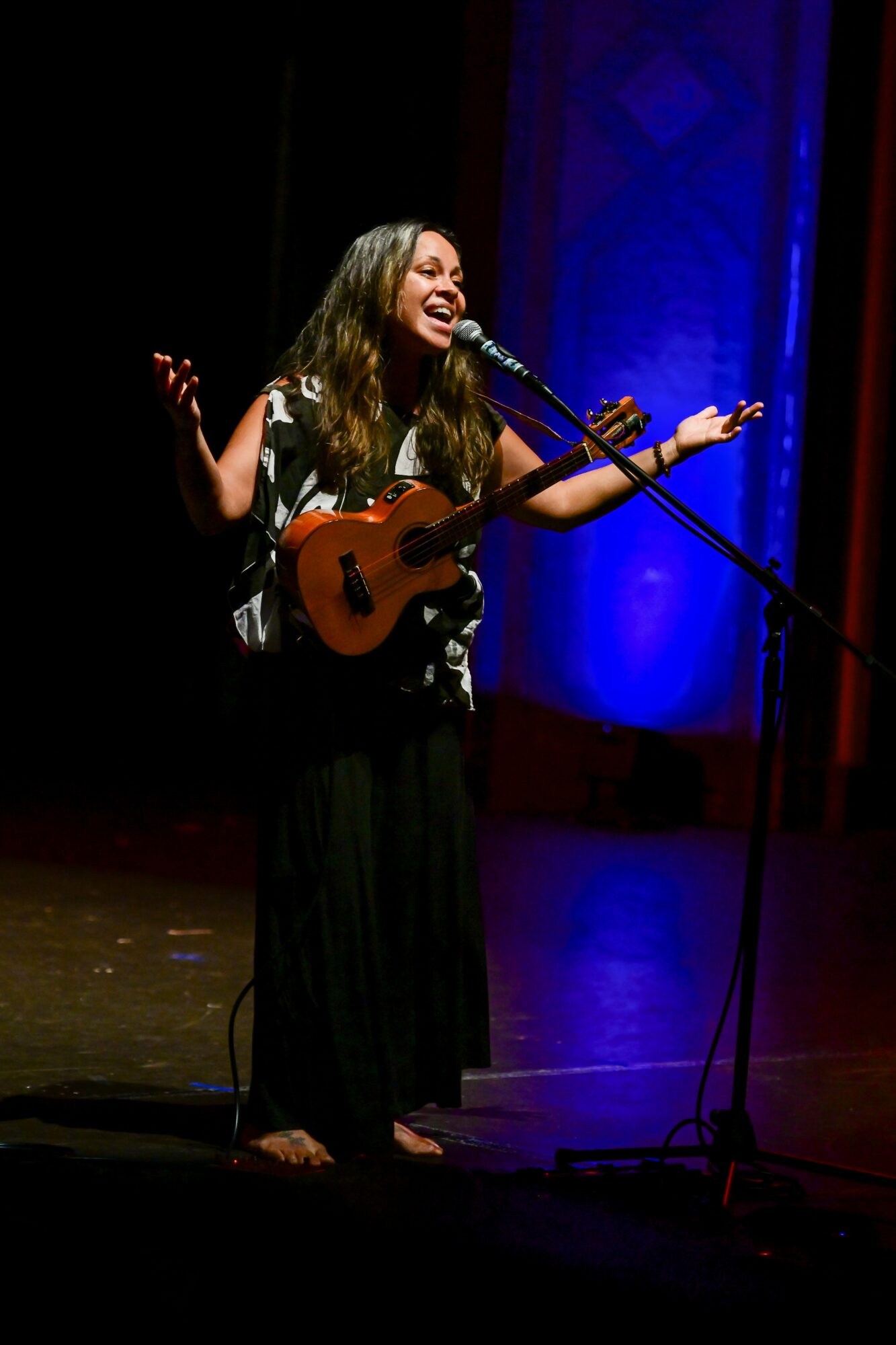 Woman singing and playing guitar on stage with blue background, smiling, with arms raised, microphone in front.