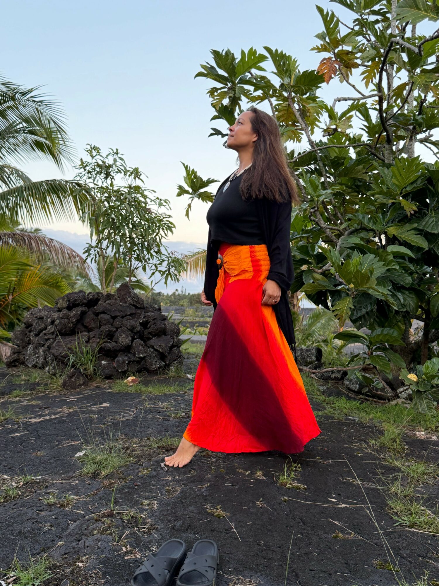 Woman in black top and red skirt walking barefoot on stone path with greenery and trees around.