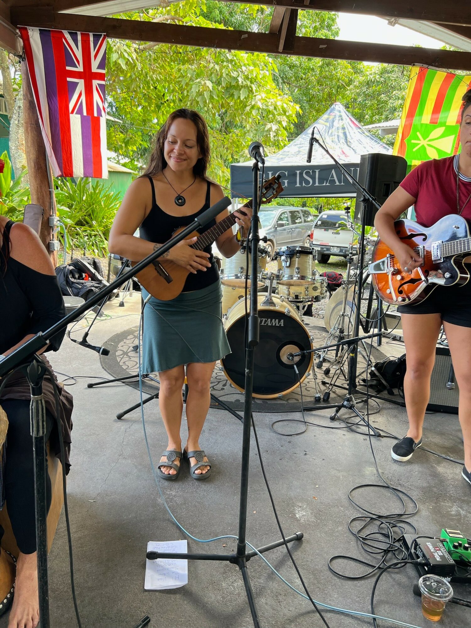 Two women playing guitars on stage with flags hanging above, green trees in background, and musical equipment around.