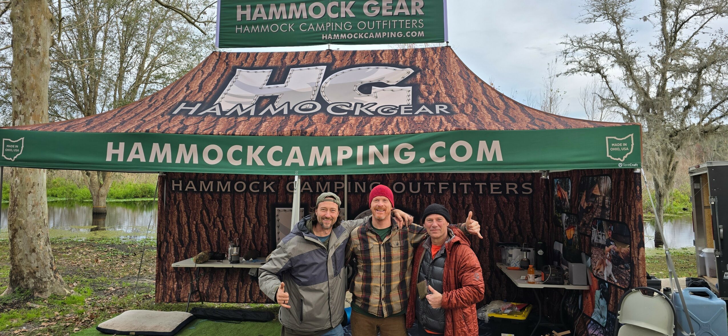 Three people stand in front of a camping gear booth with banners and trees in the background.