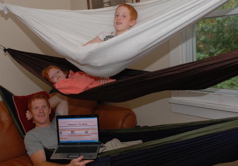Three people are lying in hammocks indoors, with a window showing trees outside. One person is using a laptop.