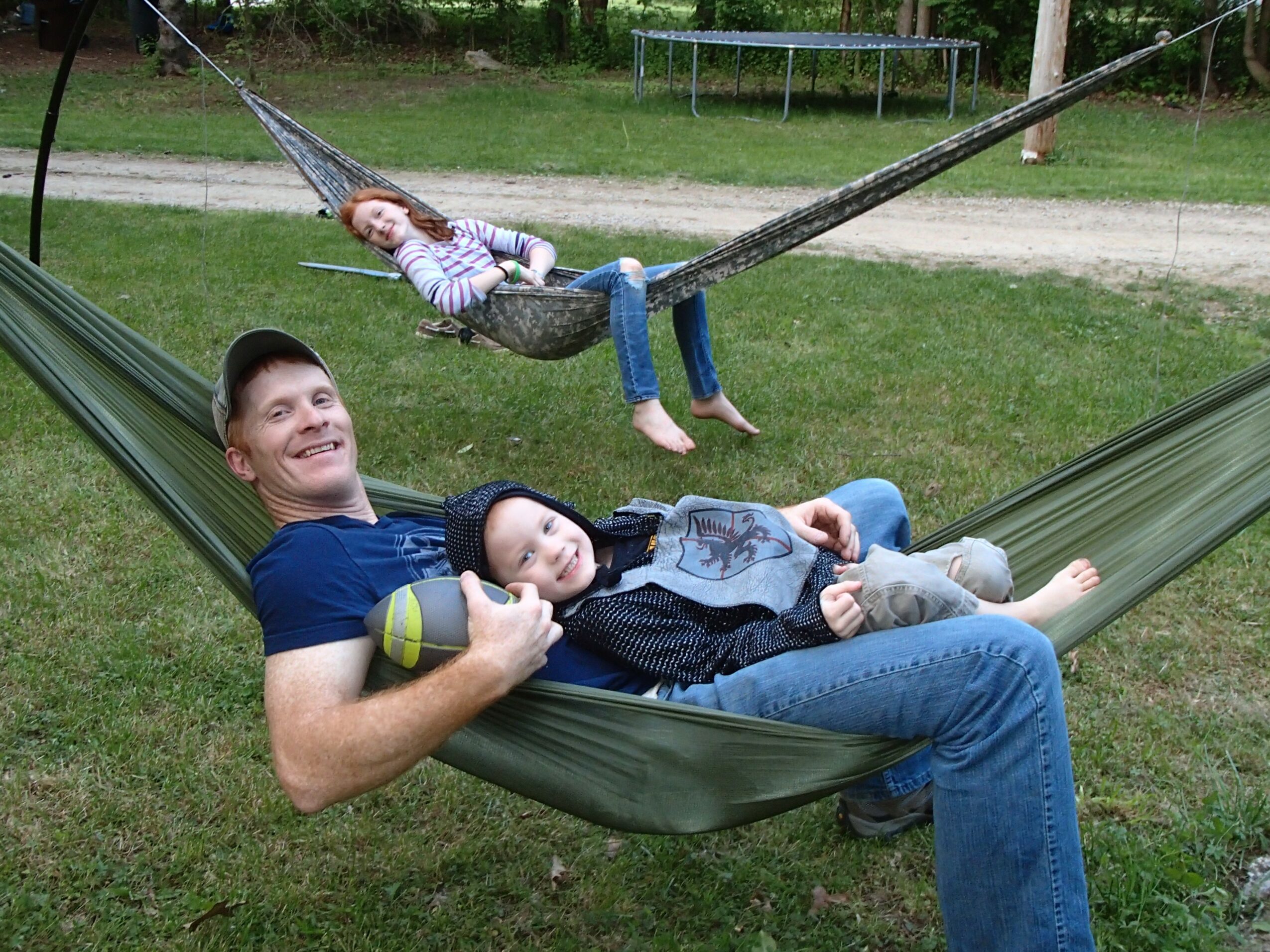 Two children and an adult relaxing on a hammock outdoors, with a girl in a swing above them, in a park setting.