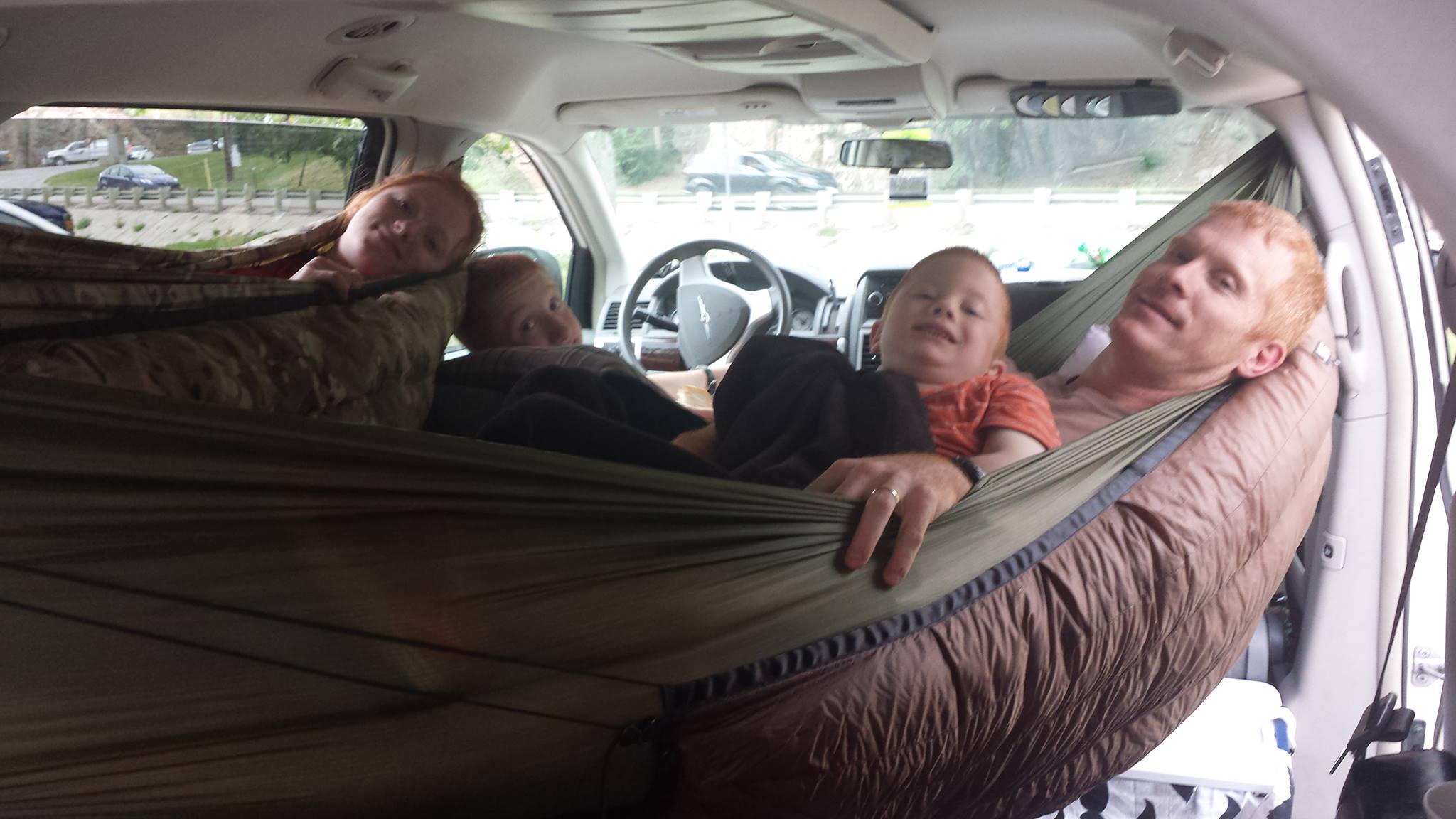 Inside a vehicle with four people lying on a hammock-like bed across the front seats, looking at the camera.