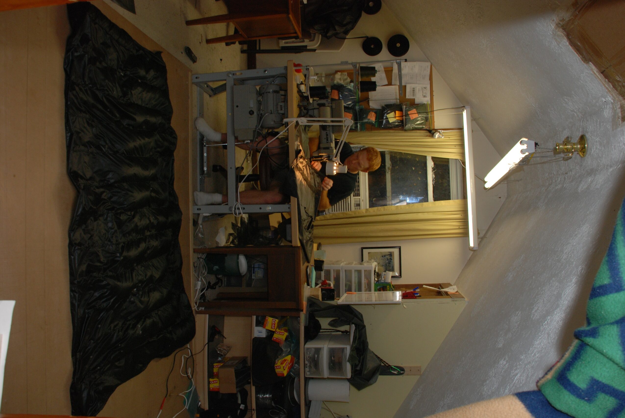 Person sitting at a desk in a room with shelves, a window, and various objects, viewed from the side.