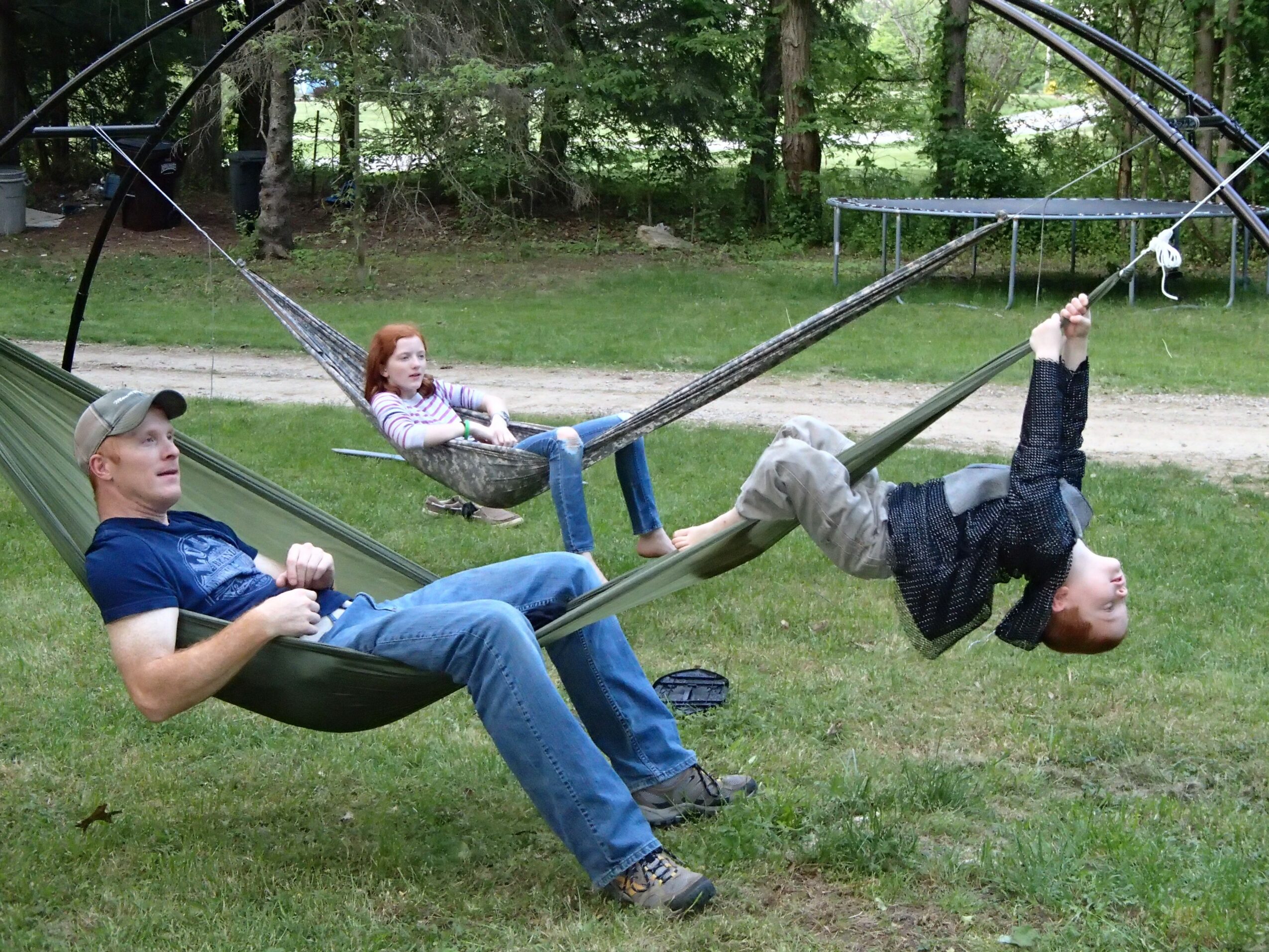 Two children and an adult on a swing set in a park, with trees and a pathway in the background.