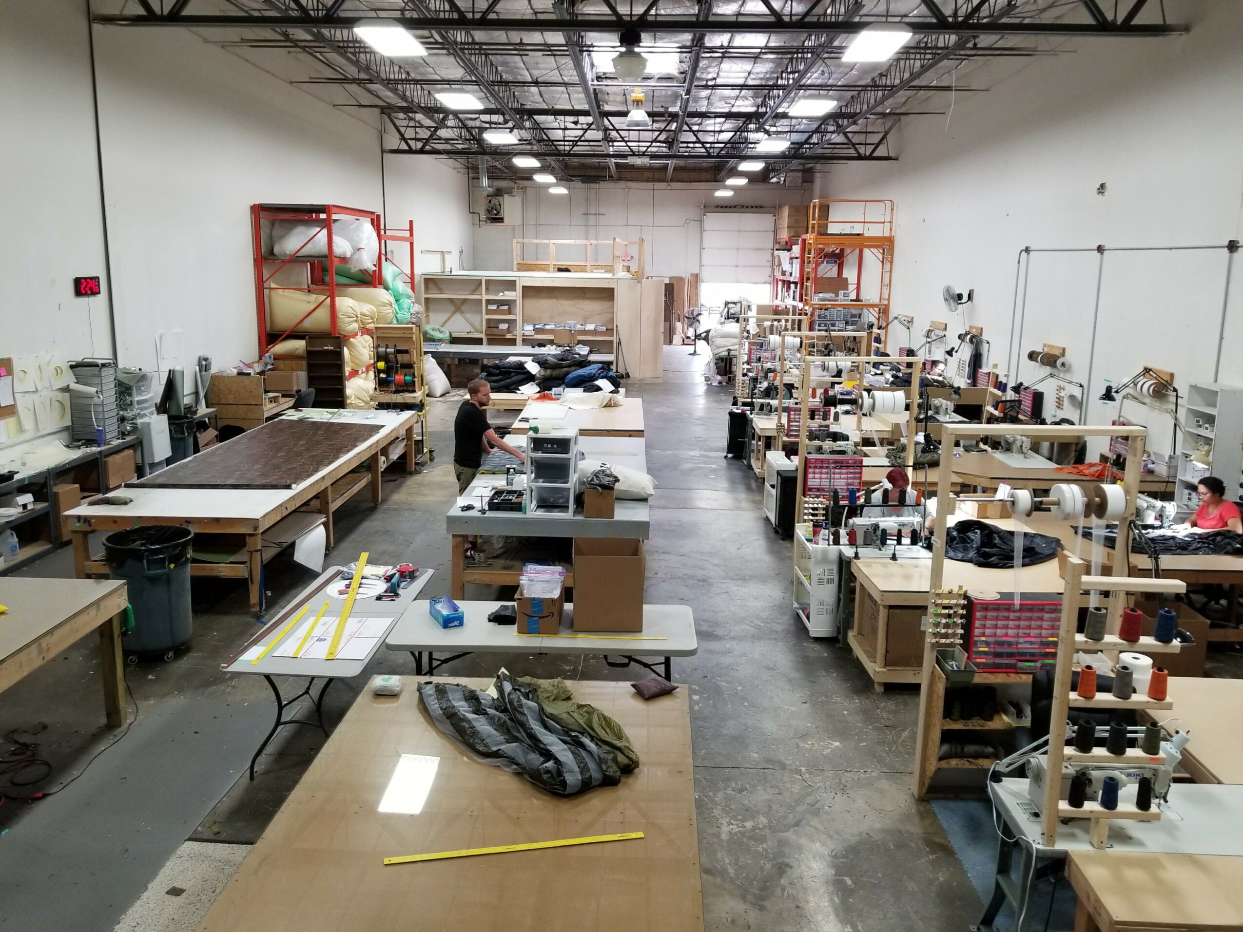 Workshop with workbenches, shelves, and people working, viewed from the front, with a high ceiling and industrial lighting.
