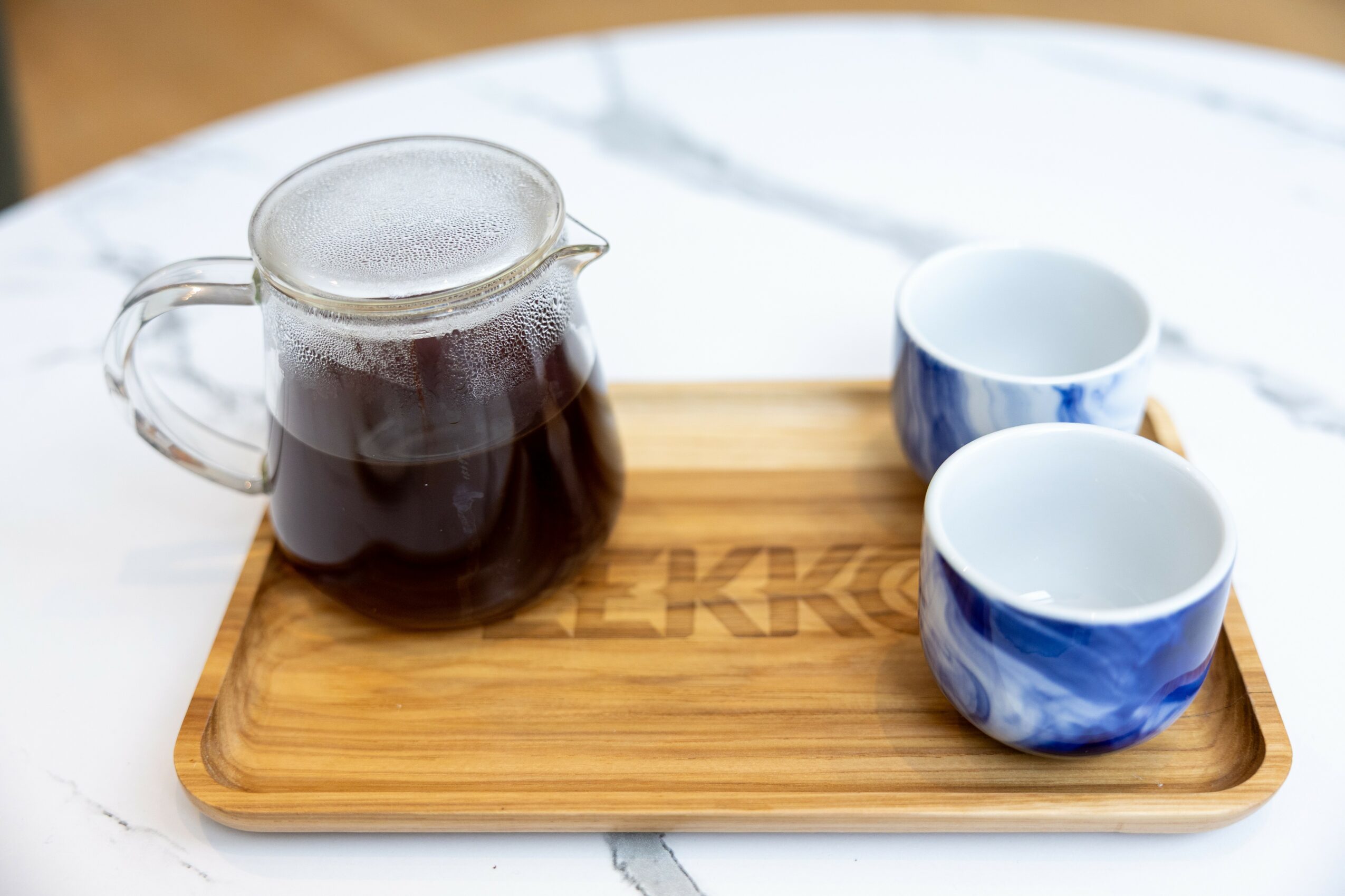 Glass pitcher with dark liquid on a wooden tray with two blue and white cups, on a white marble surface.