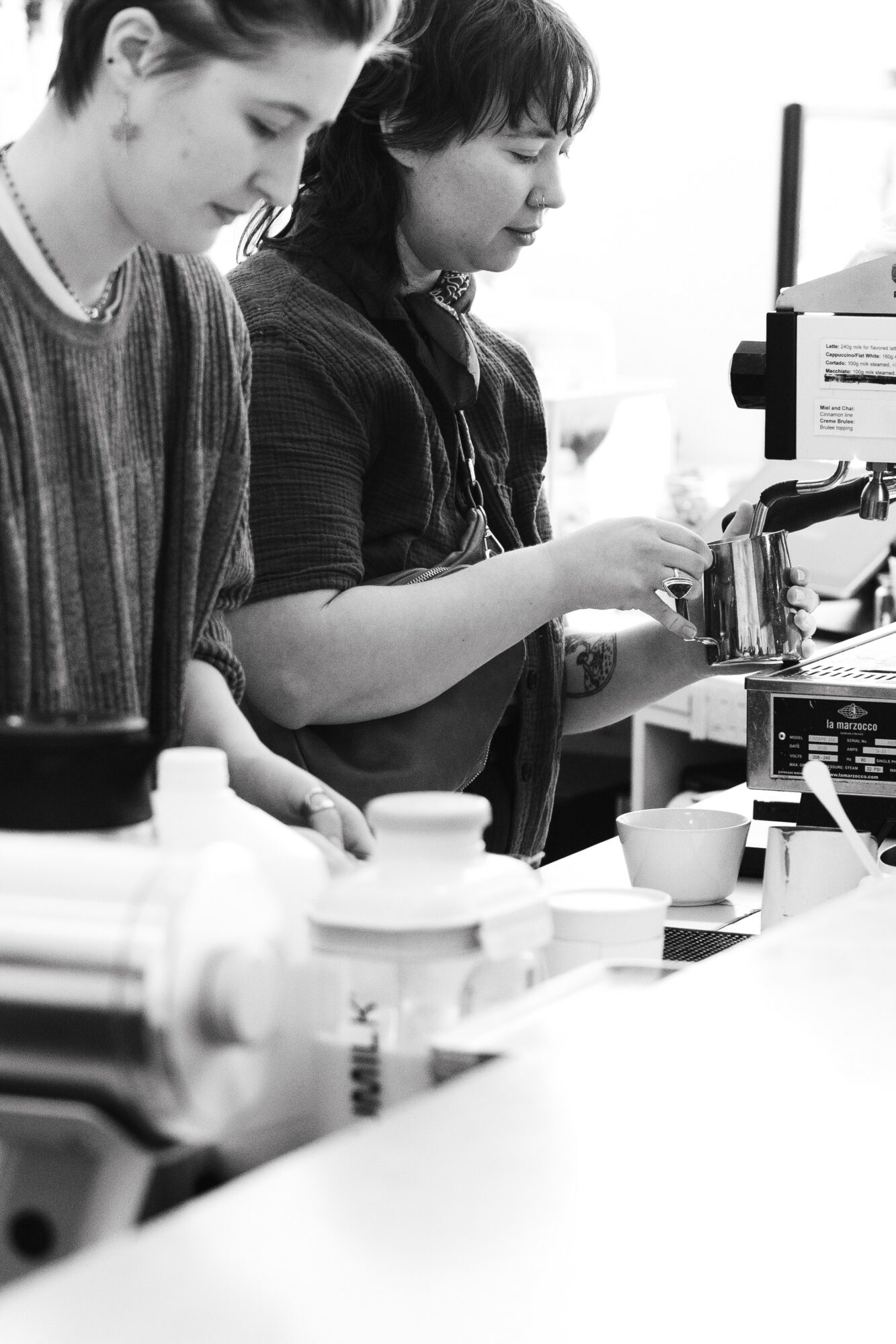 Two women working with coffee equipment in a workspace, surrounded by cups and supplies, in black and white.