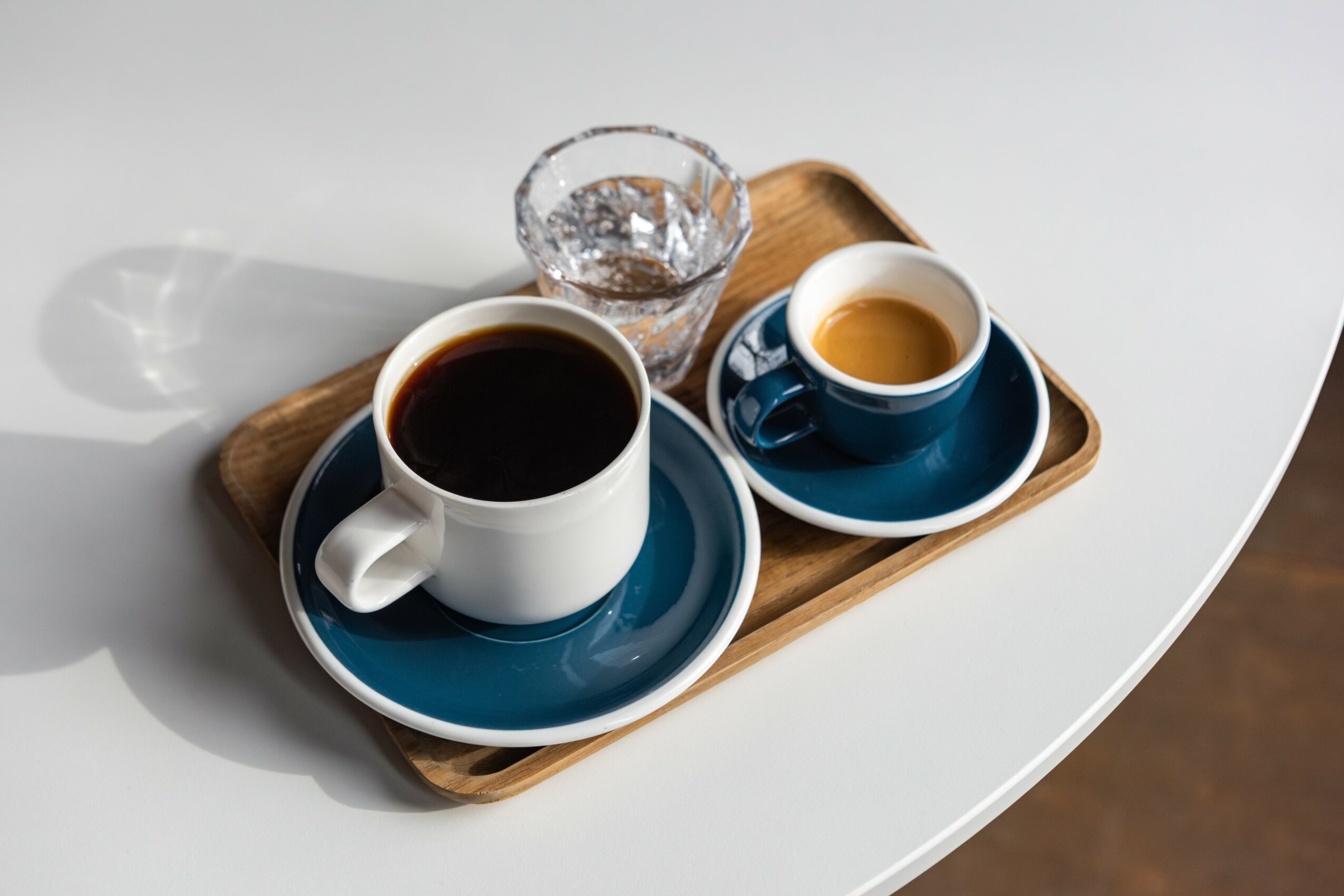 Two cups of coffee on a wooden tray with a glass of water on a white surface, with shadows, in natural light.