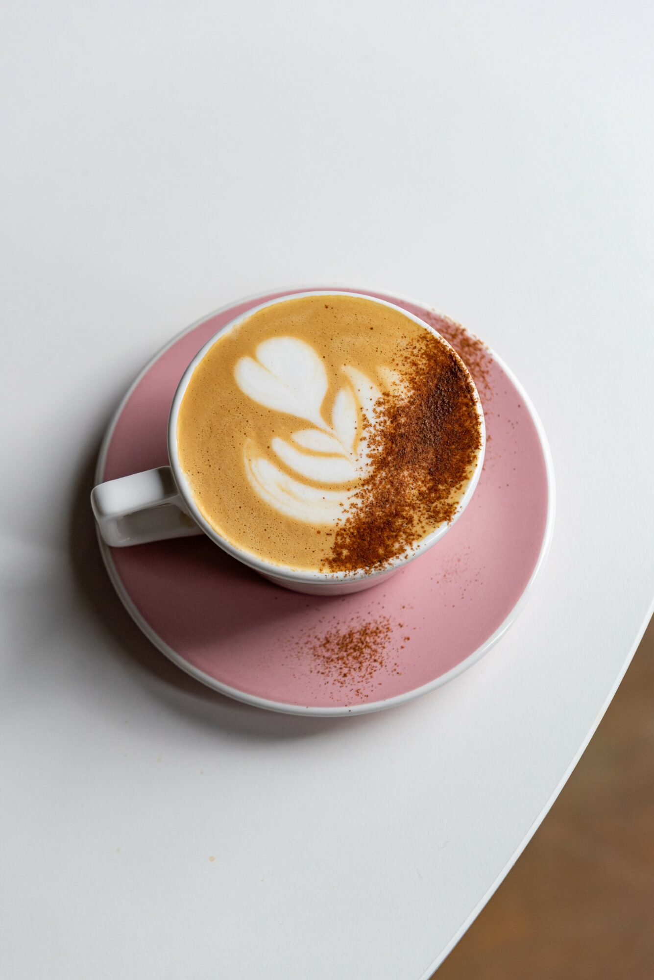 Cup of coffee with latte art on pink saucer, on white surface, with some coffee grounds on saucer and surface.