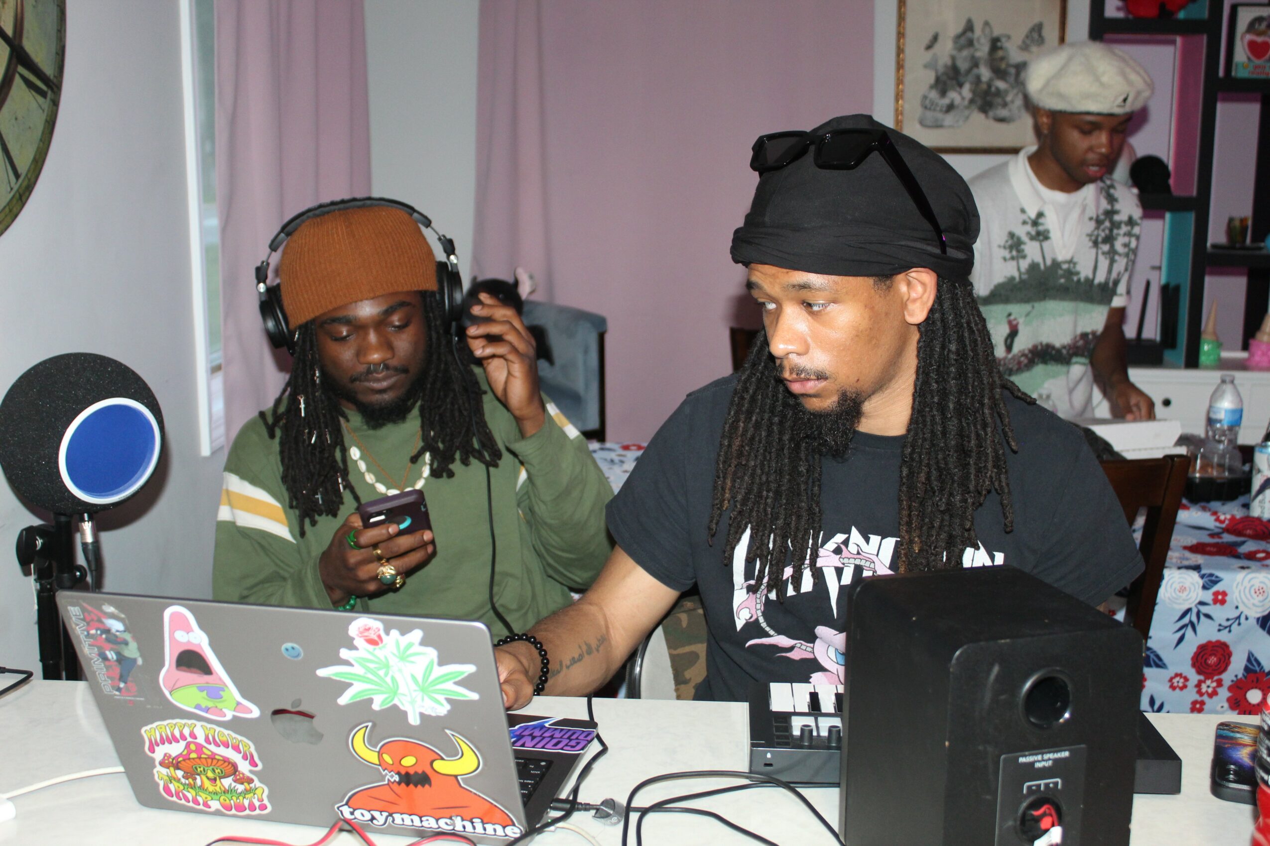 Two men working at a DJ setup with laptops, headphones, and audio equipment in a room with pink curtains and artwork.