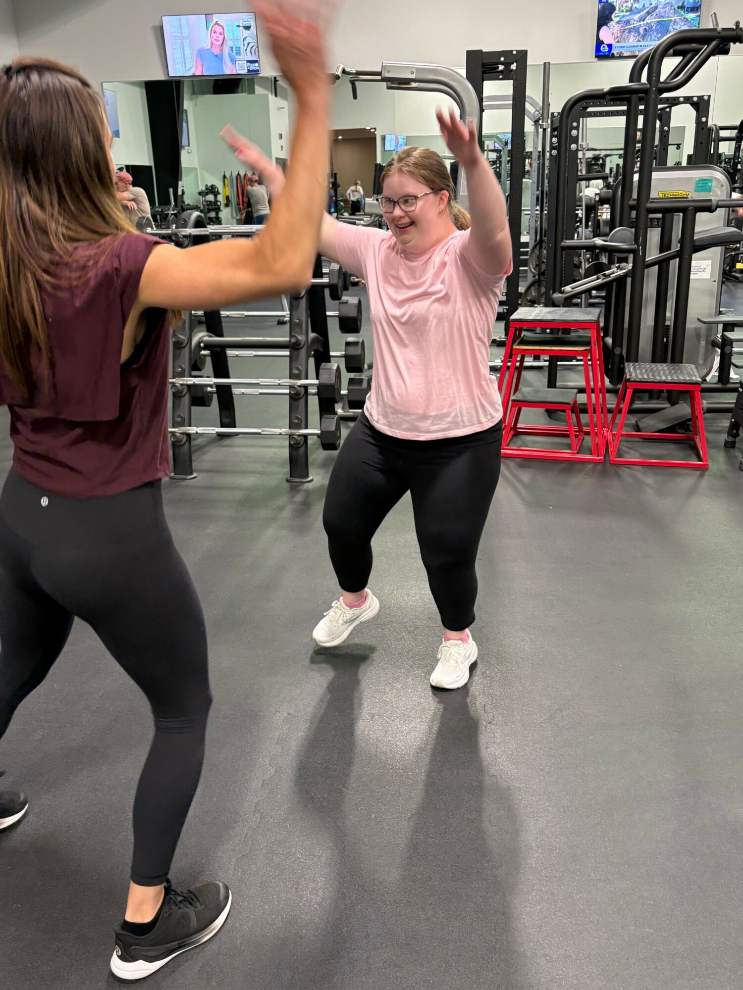 Two women in workout clothes in a gym, one stretching arm, the other standing with hands on hips, gym equipment in background.