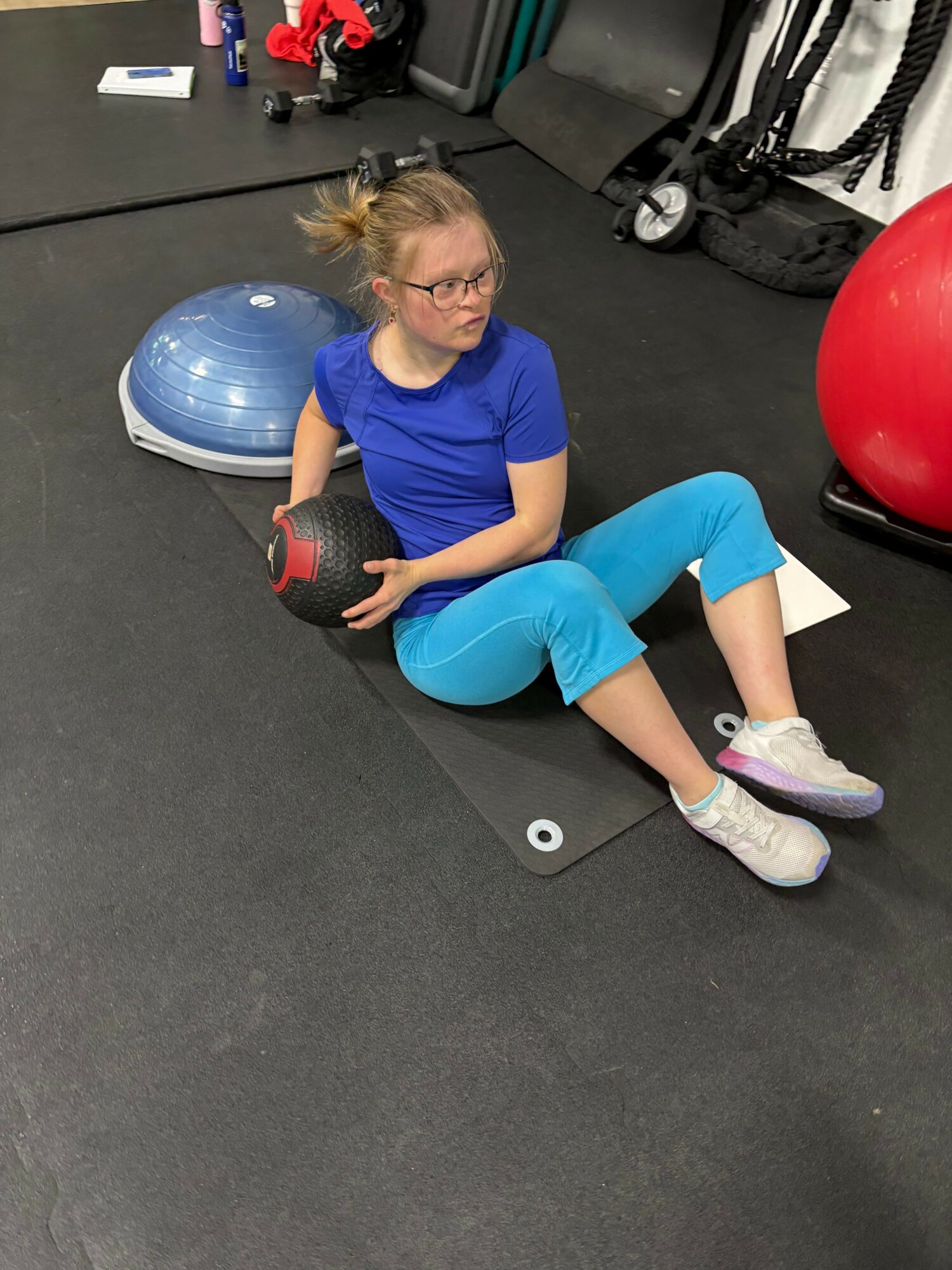 Person sitting on the floor holding a medicine ball, with a large exercise ball nearby, in a gym setting.