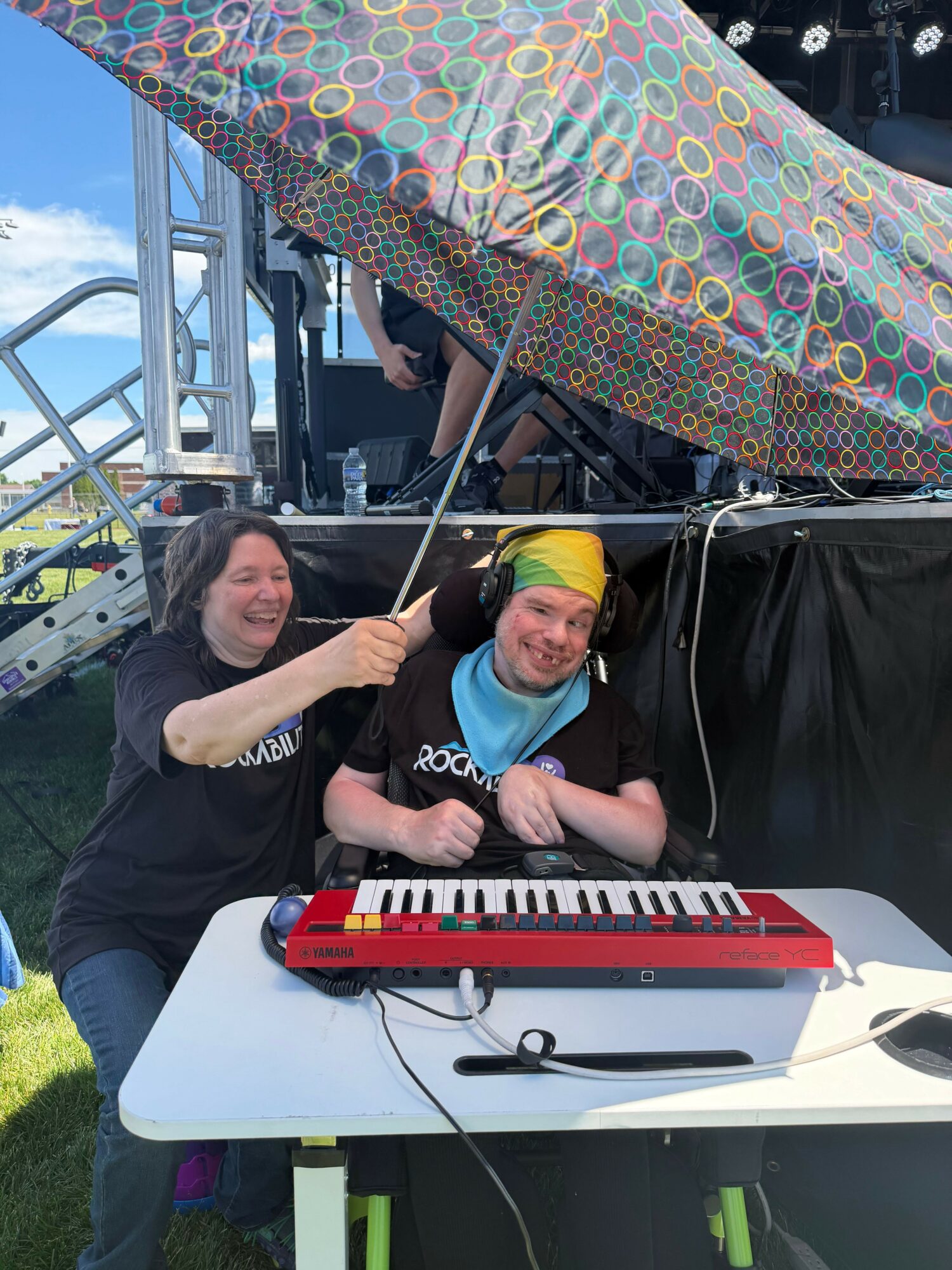 Two people sitting at a table with a red keyboard, outdoors, with a colorful tent nearby.