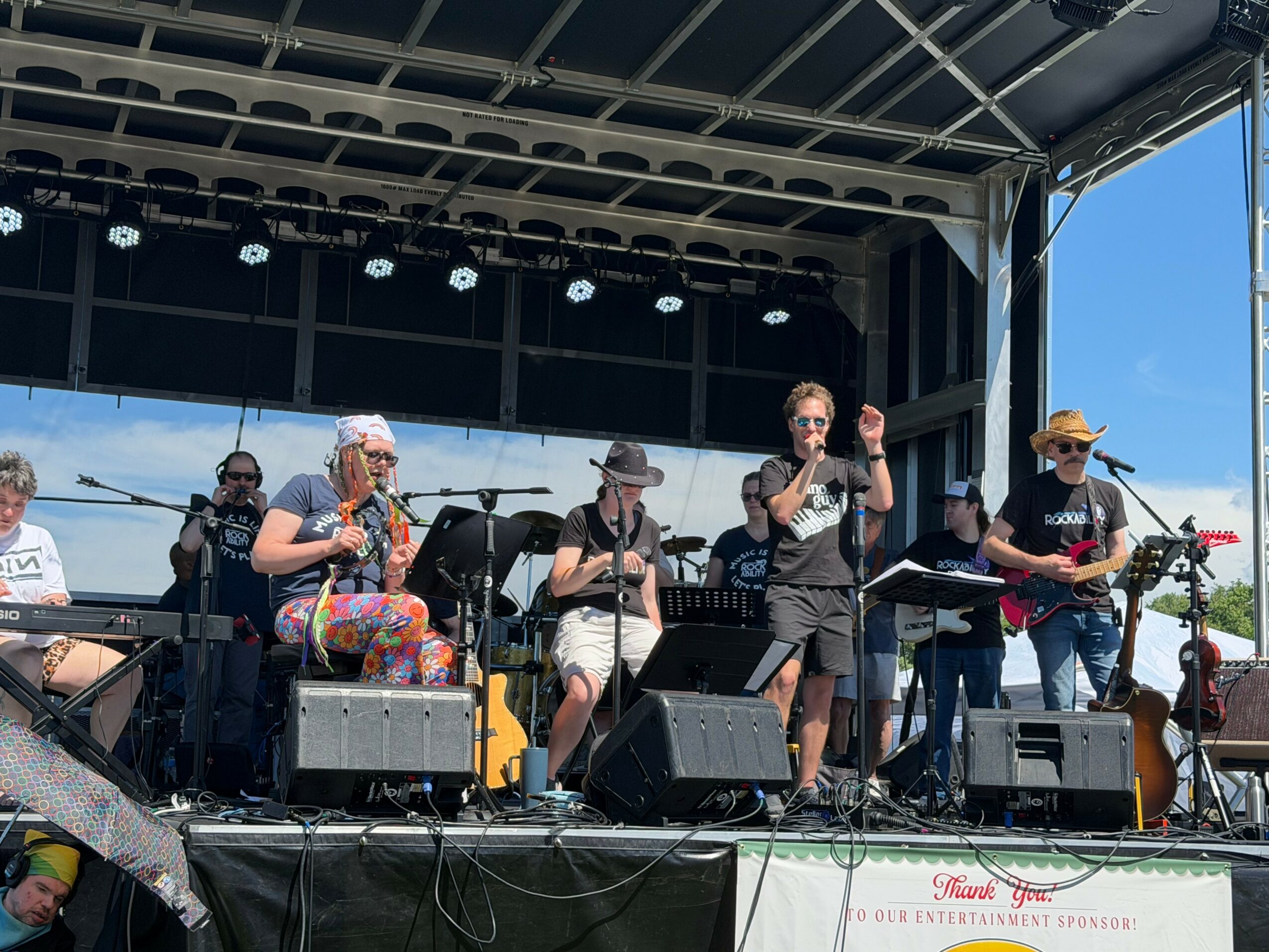 Group of musicians performing on an outdoor stage with instruments and microphones, under a metal canopy, with a blue sky background.