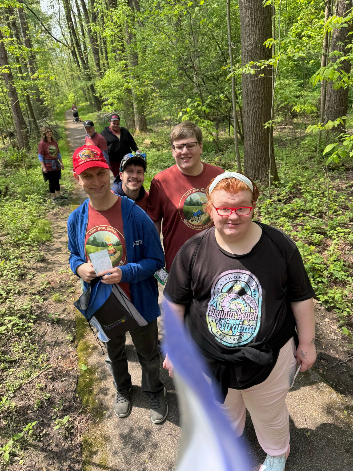 Four people sitting on a trail in a forest, smiling, with trees and greenery around them.