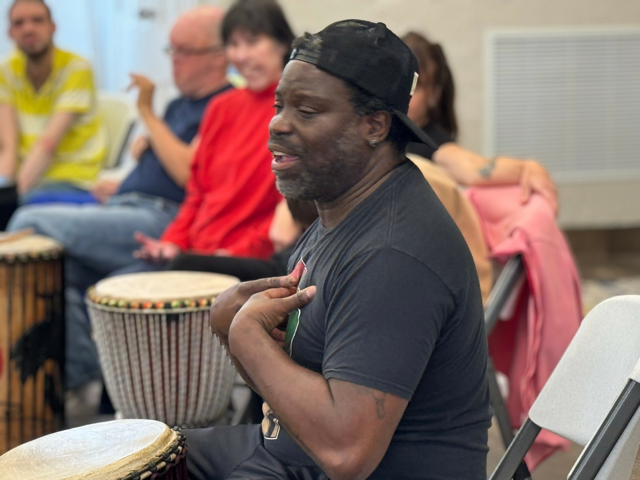 Man with a beard and cap, sitting with hand on chest, surrounded by people, some playing drums, in a casual indoor setting.