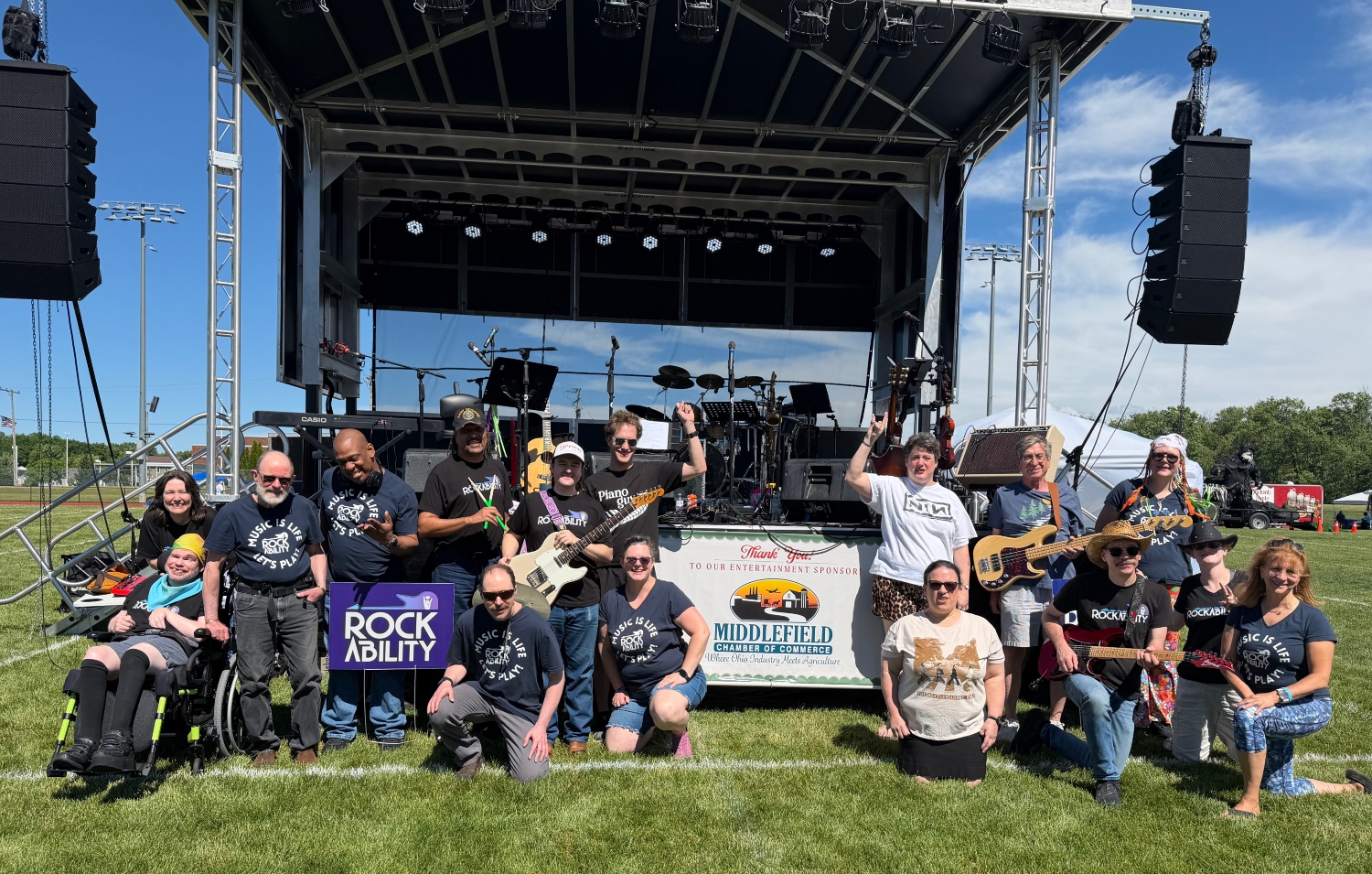 Group of people posing in front of a stage with musical instruments outdoors on grass, with a blue sky and clouds.