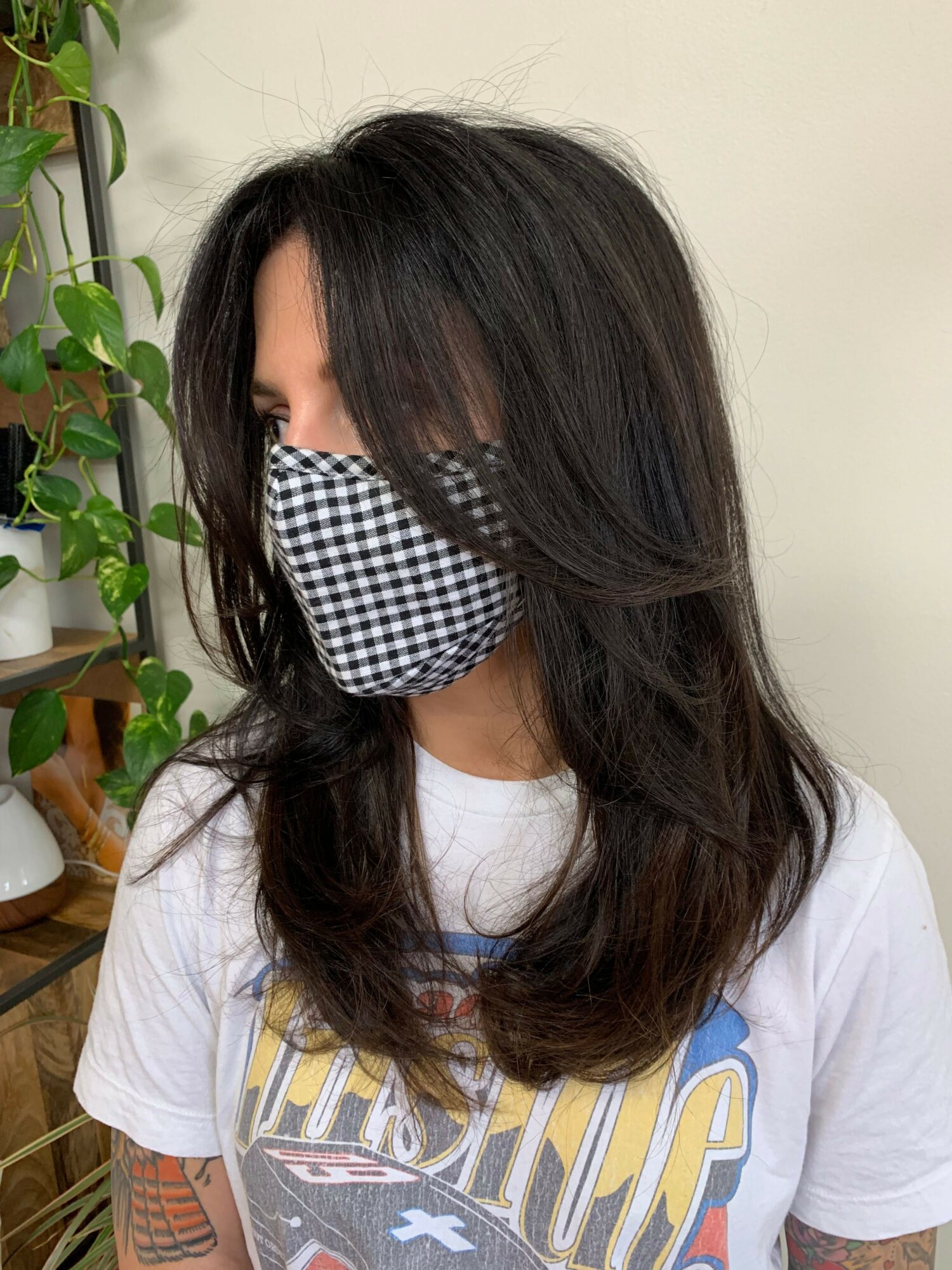 Person with long dark hair wearing a checkered face mask and a white shirt, sitting near a plant.