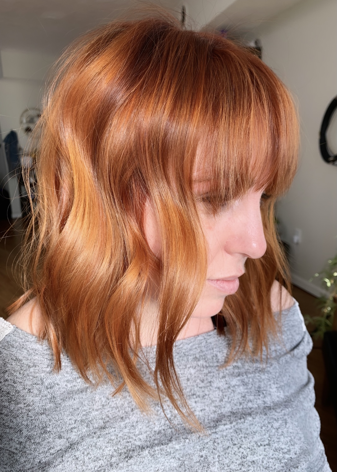 Side view of a woman with wavy, shoulder-length red hair and bangs, wearing a gray top.