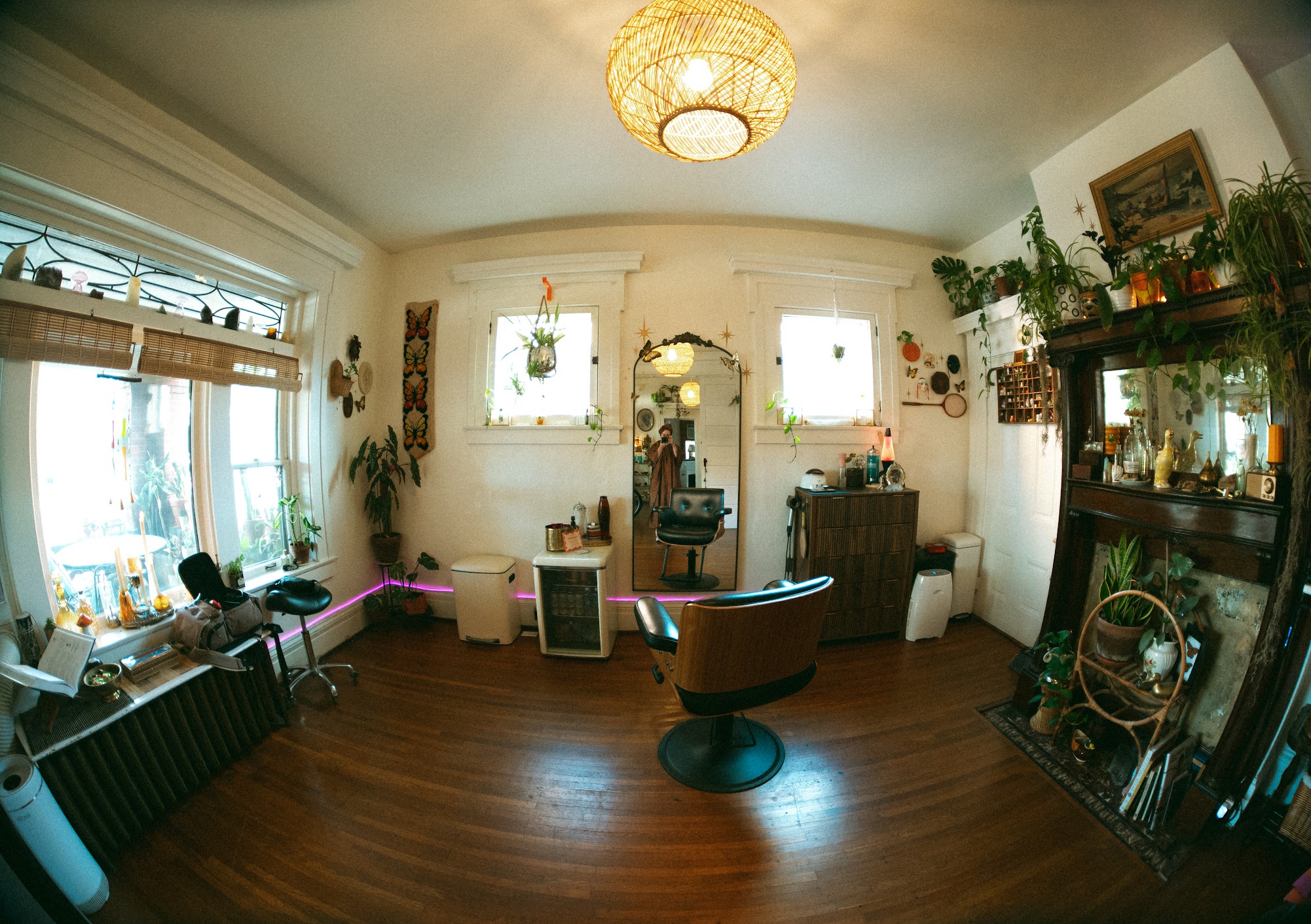 Living room with large window, plants, a chair, and a display cabinet, illuminated by a ceiling light. Wooden floor and white walls.