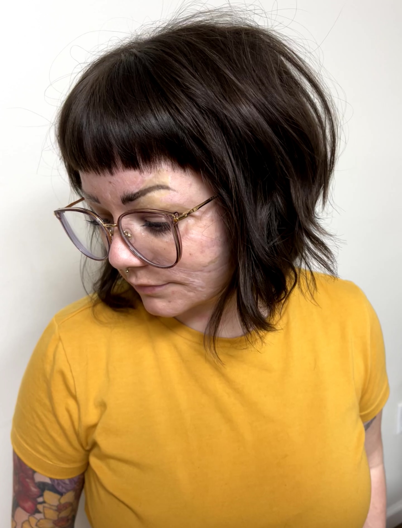 Woman with dark, layered hair and glasses wearing a yellow shirt, looking downward, against a plain background.