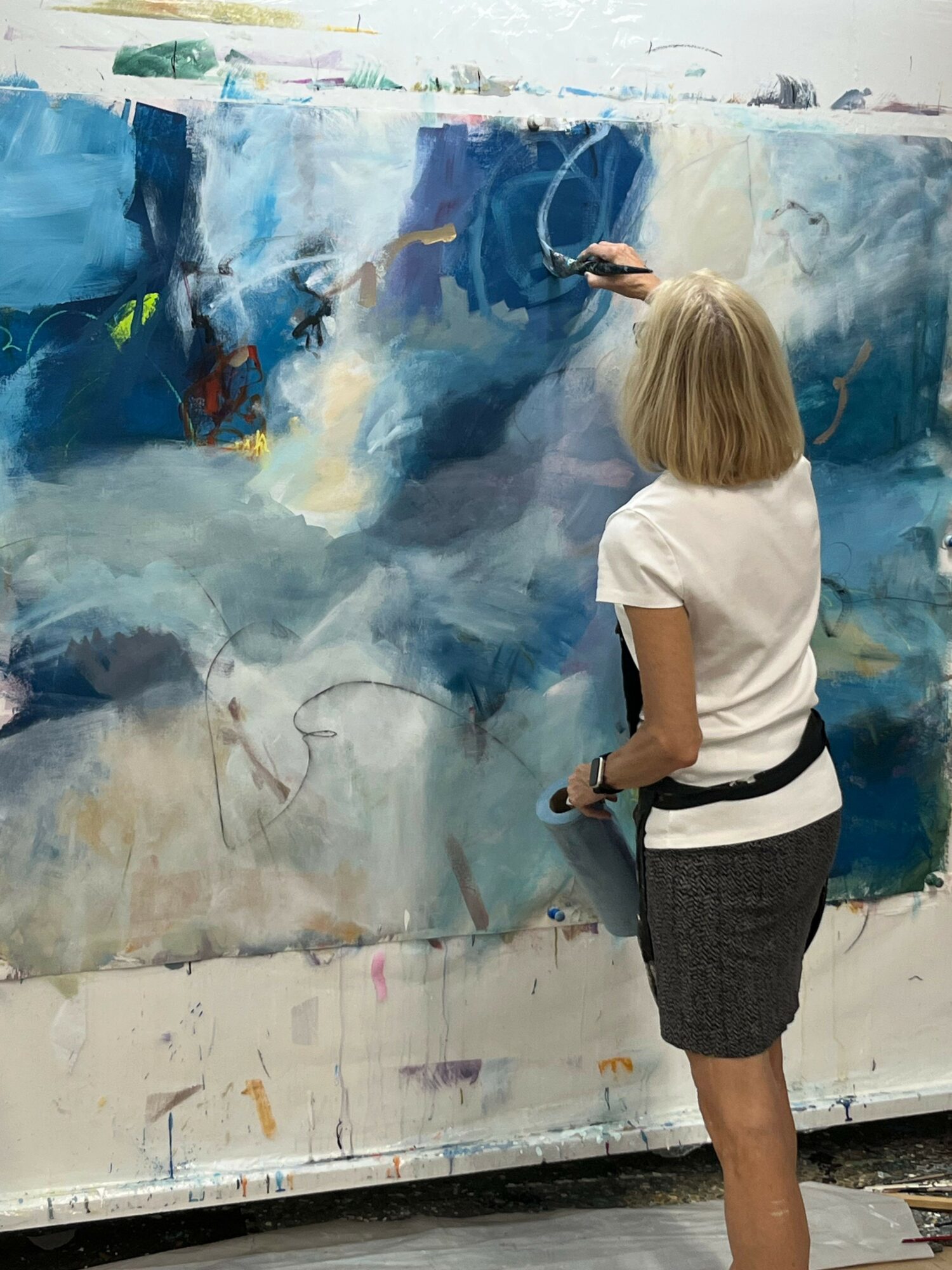 Person with blonde hair painting on a large canvas, wearing a white shirt and dark skirt, facing the artwork.
